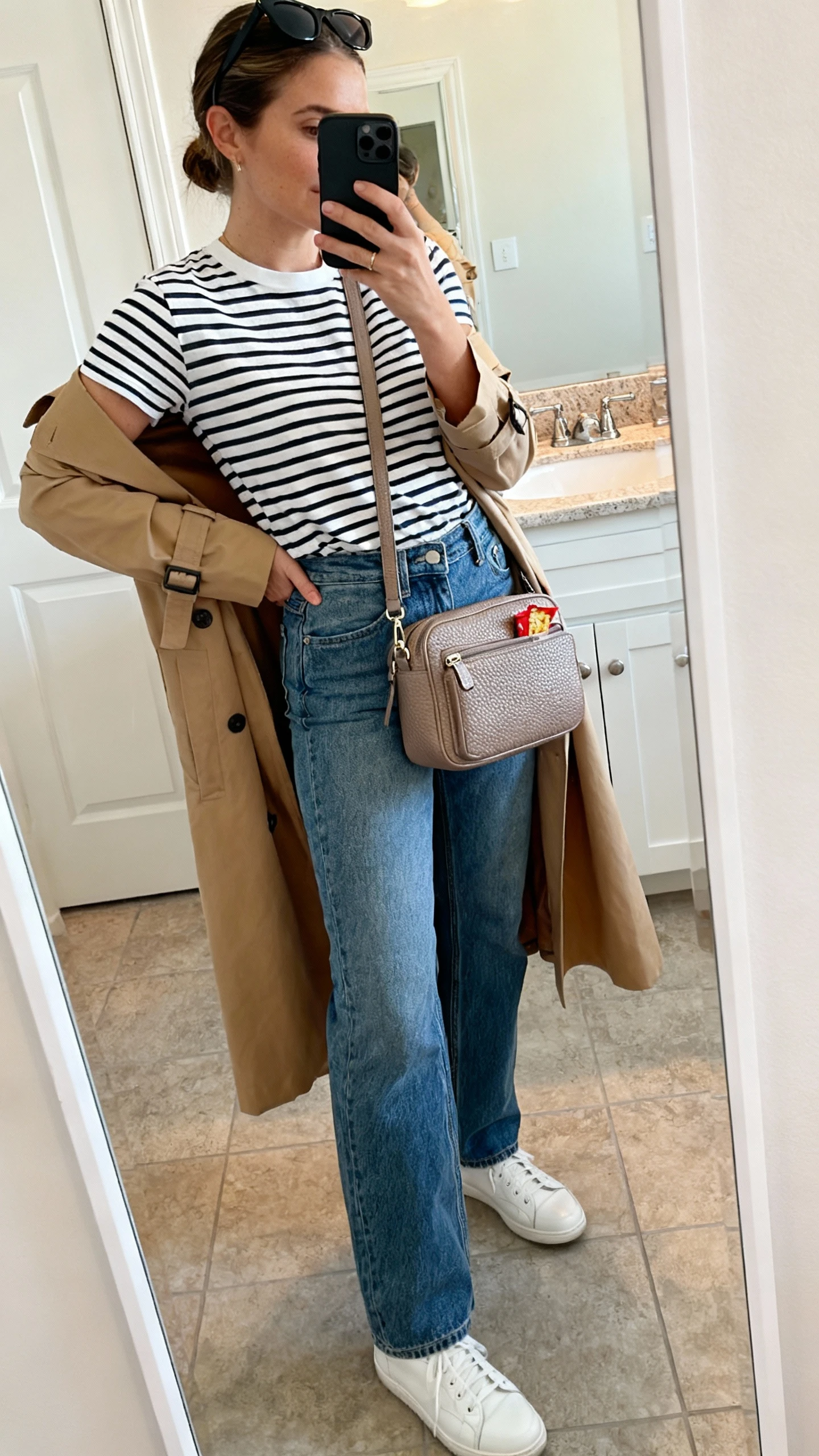 iPhone side-angle mirror selfie of a woman wearing a striped Breton tee tucked into high-rise straight-leg jeans, tan trench coat draped open, white leather sneakers, a roomy taupe pebbled-leather crossbody with snack-sized compartments, and black sunglasses perched on her head, face not visible, bathroom mirror, soft indoor lighting, iPhone photo quality