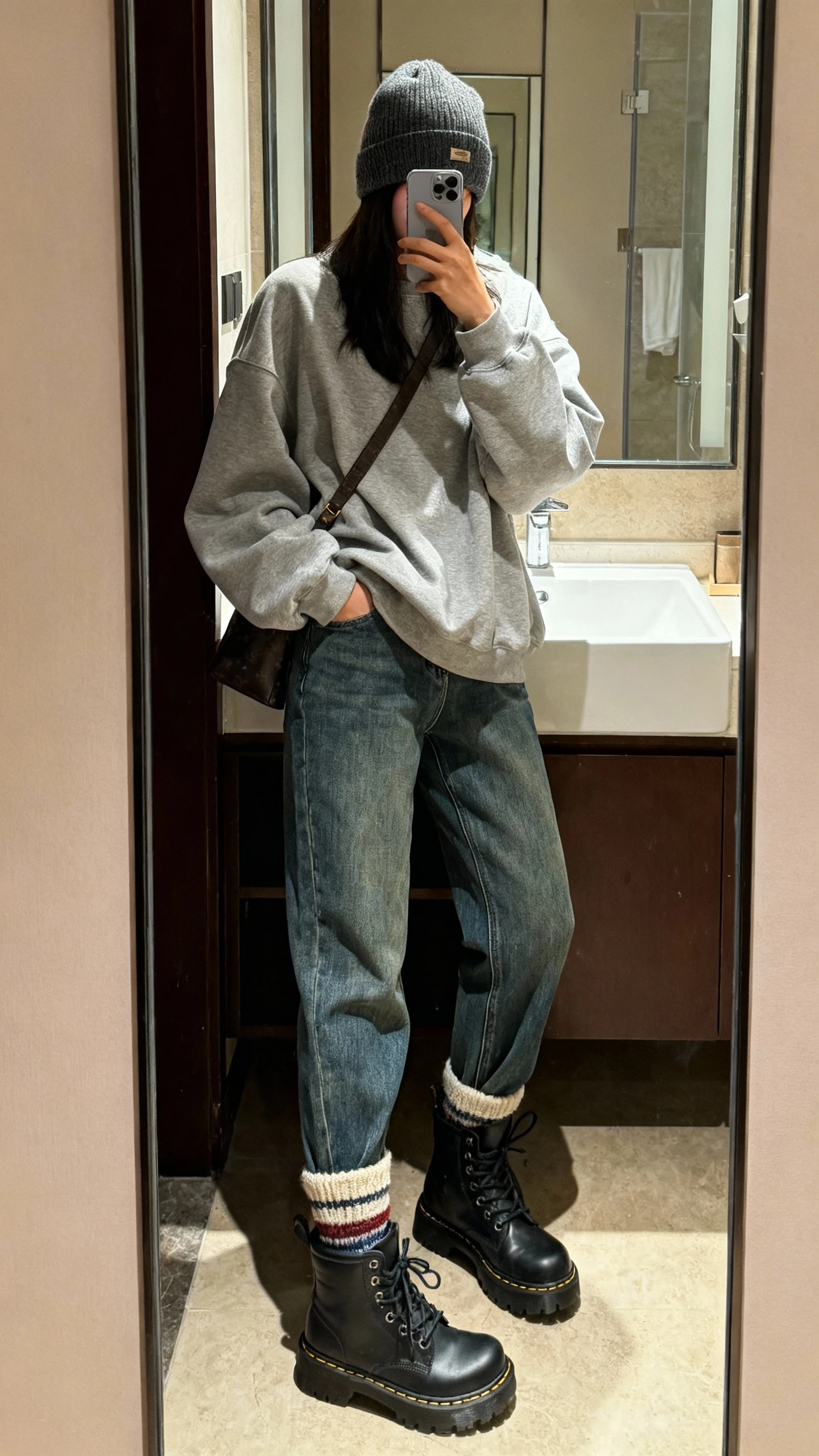 iPhone side-angle mirror selfie of a woman wearing relaxed straight-leg jeans, an oversized sweatshirt, chunky lace-up boots, cozy crew socks peeking above the boots, a beanie and crossbody bag, face not visible, bathroom mirror, warm indoor lighting, iPhone photo quality.