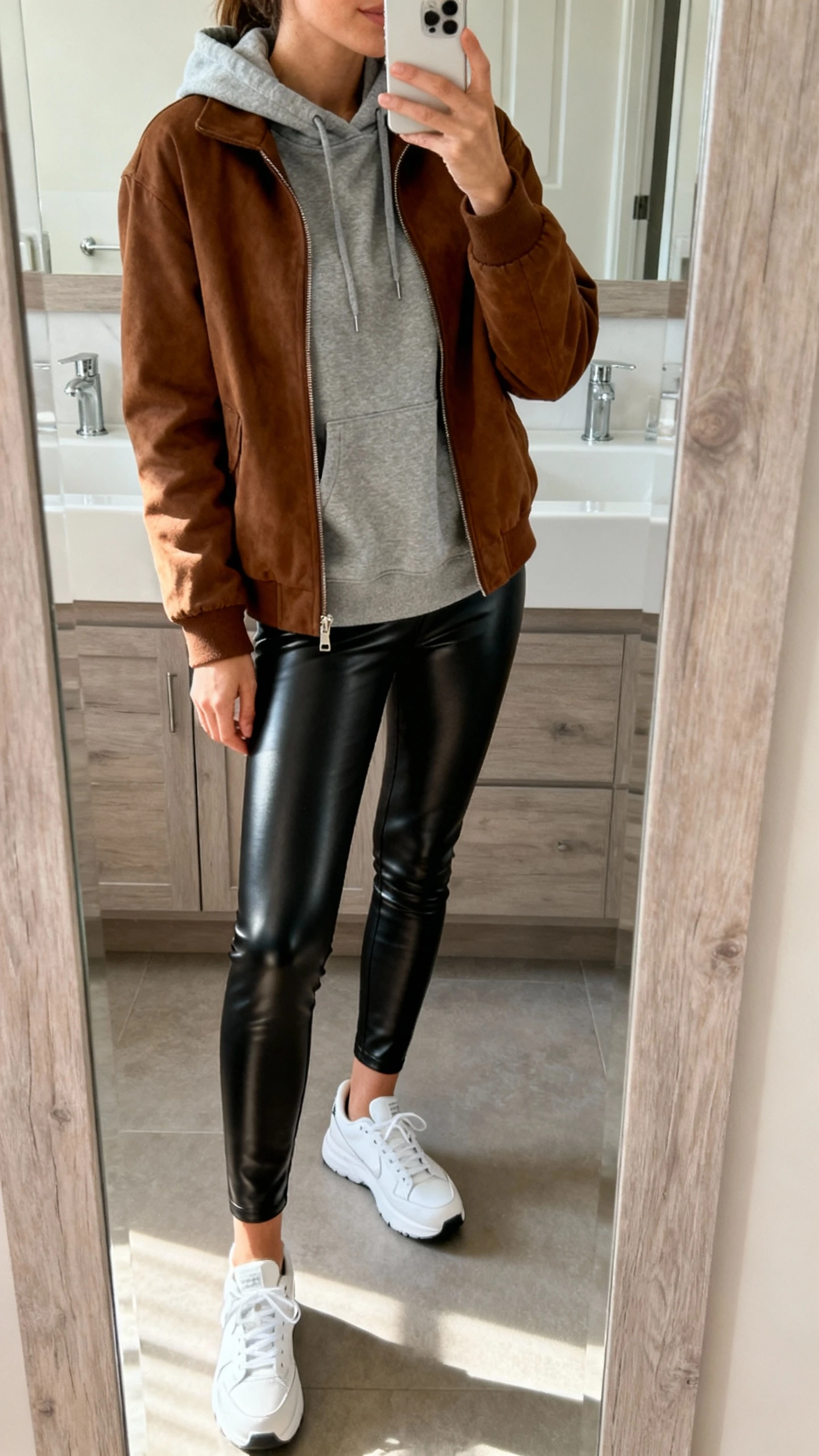 iPhone side-angle mirror selfie of a woman wearing a brown jacket over a gray hoodie, black leather leggings, and sleek white sneakers, face not visible, bathroom mirror, soft indoor morning light, iPhone photo quality.