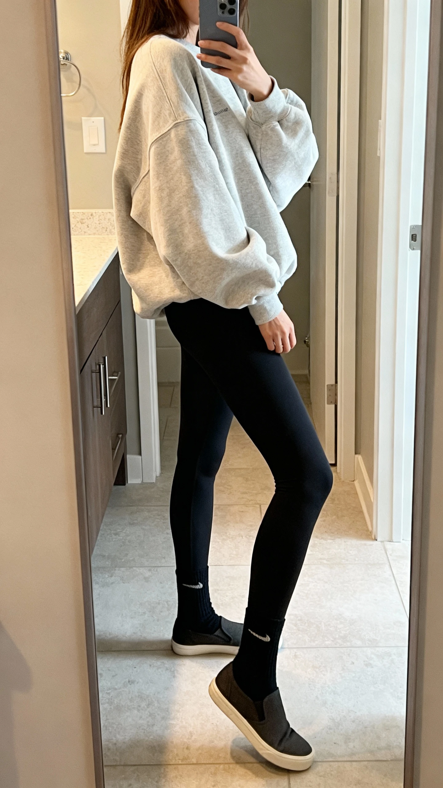 iPhone side-angle mirror selfie of a woman wearing an oversized sweatshirt, sleek black leggings, ankle socks, and minimalist sneakers, face not visible, bathroom full-length mirror, soft indoor lighting, iPhone photo quality.