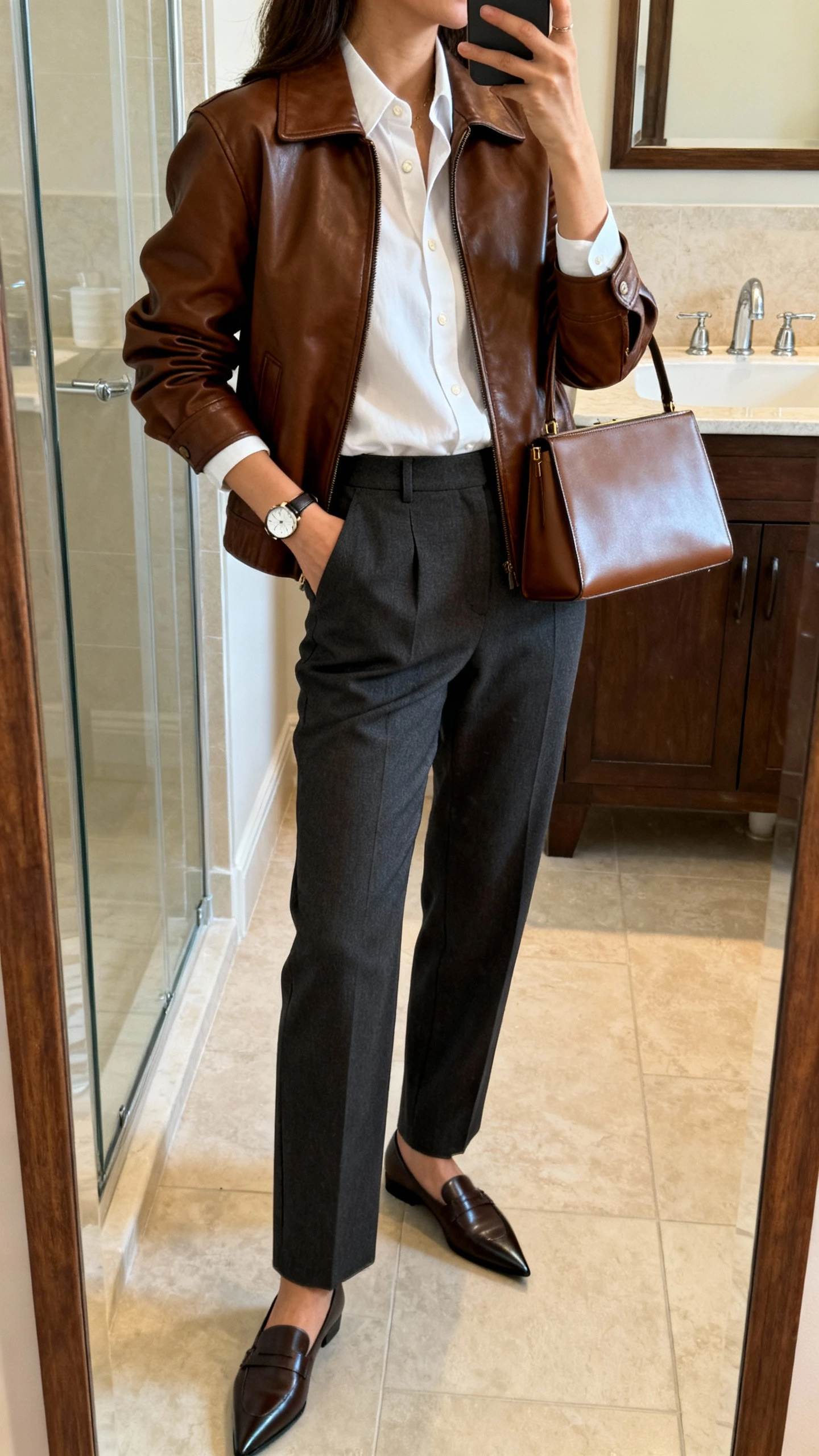 iPhone side-angle mirror selfie of a woman wearing a brown leather jacket layered over a crisp white button-down, tailored charcoal trousers, pointed-toe loafers, slim leather tote, subtle watch, face not visible, bathroom full-length mirror, morning indoor lighting, iPhone photo quality