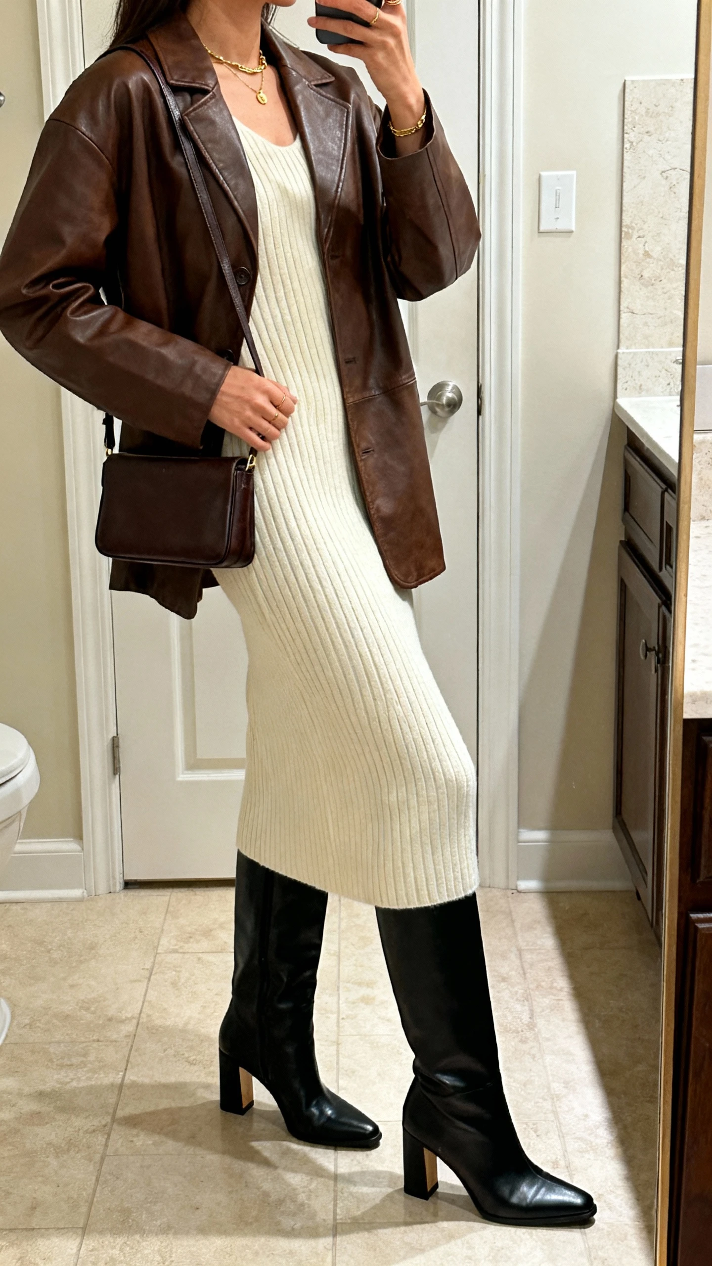 iPhone side-angle mirror selfie of a woman wearing a brown leather jacket layered over a ribbed cream knit midi dress, black knee-high leather boots with a structured heel, a slim leather shoulder bag, and delicate gold jewelry; face not visible, bathroom mirror, warm indoor lighting, iPhone photo quality.