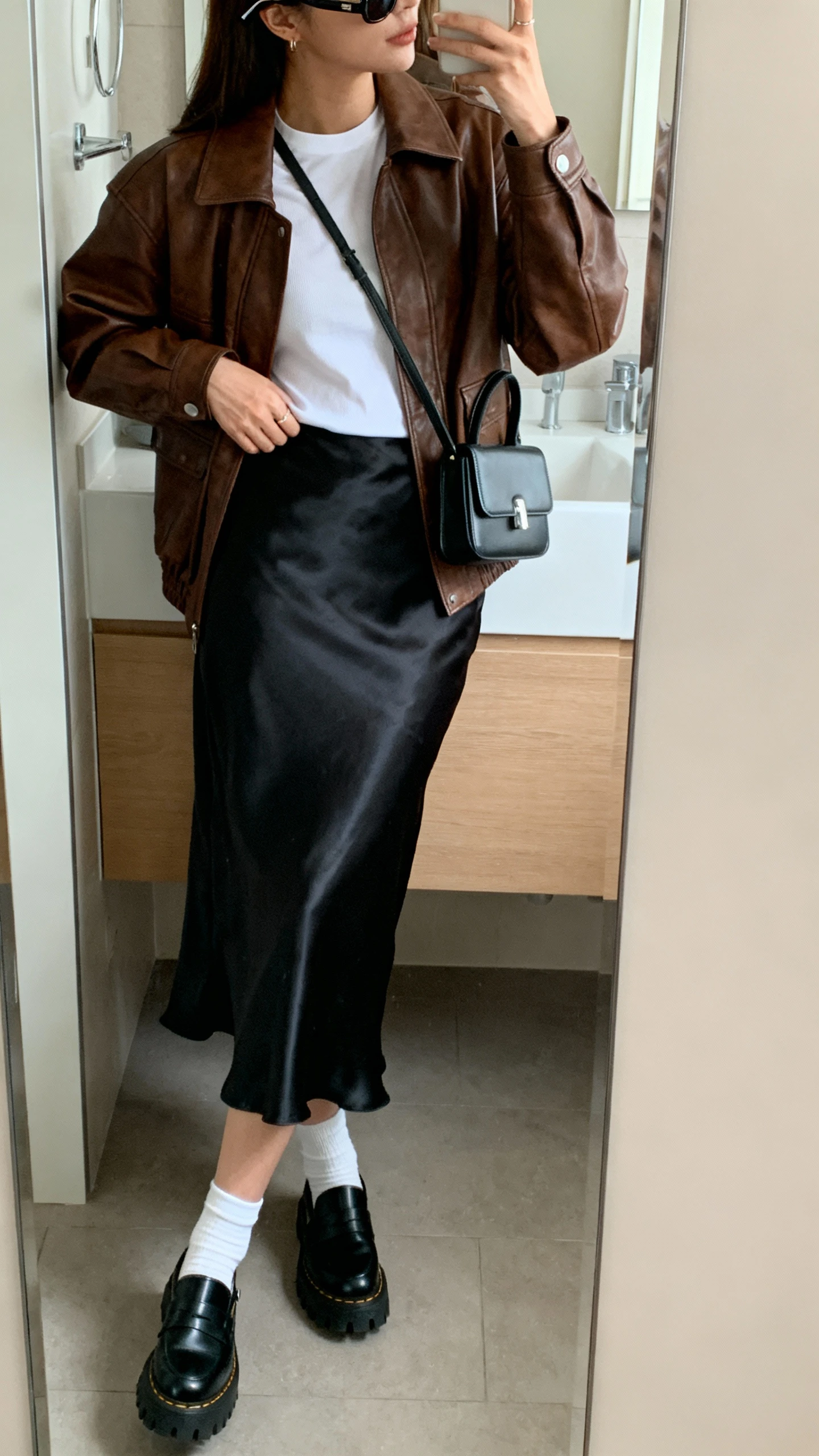 iPhone side-angle mirror selfie of a woman wearing a brown leather jacket over a black satin slip skirt, white fitted tee, black lug-sole loafers with white socks, structured mini shoulder bag, and slim sunglasses pushed up on head, face not visible, bathroom mirror, soft morning light, iPhone photo quality.