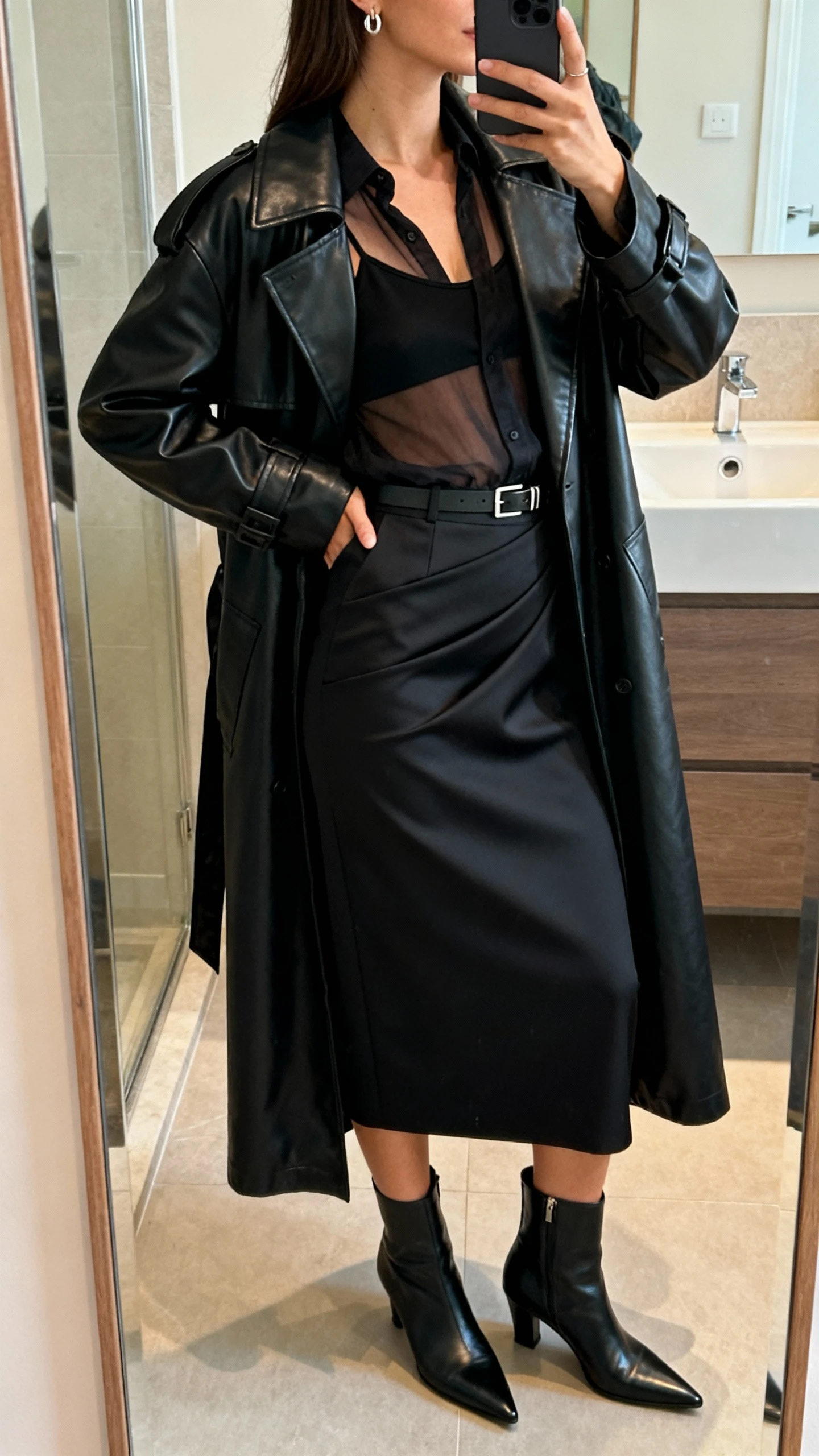 iPhone side-angle mirror selfie of a woman wearing a long black leather trench over a sheer black blouse layered with a sleek camisole, bias-cut midi skirt, and pointed ankle boots, subtle earrings and a slim belt, face not visible, bathroom mirror, soft indoor lighting, iPhone photo quality.