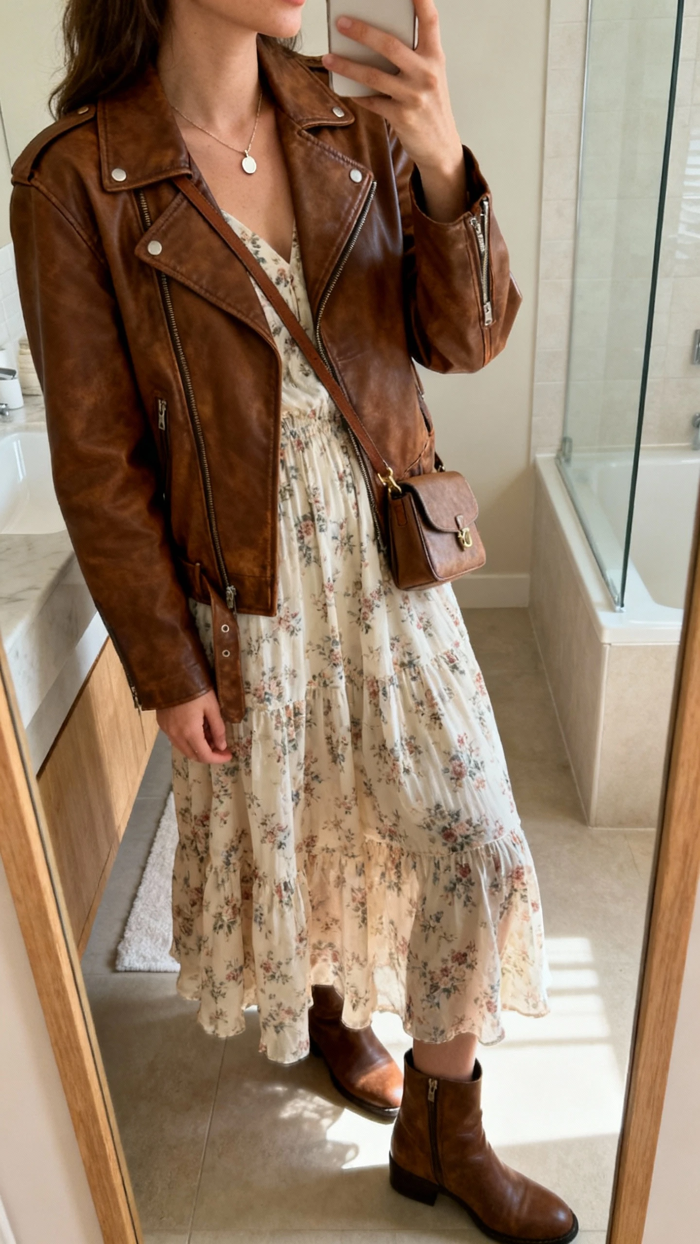 iPhone side-angle mirror selfie of a woman wearing a retro brown leather jacket over a floaty floral midi dress, ankle boots, delicate pendant necklace, and small crossbody bag, face not visible, bathroom mirror, morning light, iPhone photo quality.