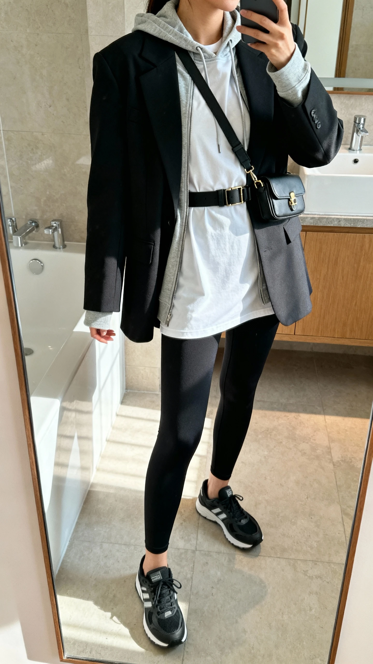 iPhone side-angle mirror selfie of a woman wearing elevated athleisure: high-waist black leggings, longline tee, lightweight hoodie layered under a structured blazer, sleek sneakers, crossbody belt bag, face not visible, bathroom mirror, indoor daylight, iPhone photo quality.