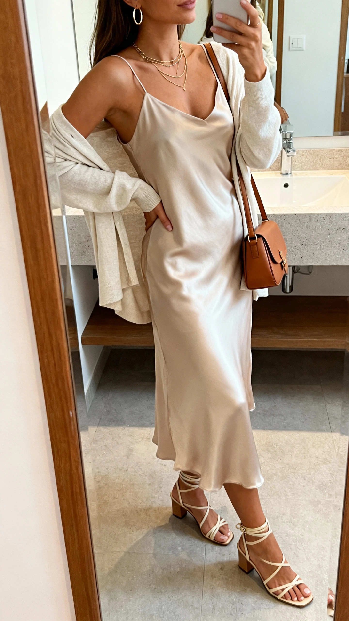 iPhone side-angle mirror selfie of a woman wearing the piazza-to-dinner slip dress formula: midi silk slip dress, light cardigan draped over shoulders, strappy low-heel sandals, small leather shoulder bag, delicate layered necklace, and hoop earrings; face not visible, bathroom mirror with soft indoor lighting, iPhone photo quality.
