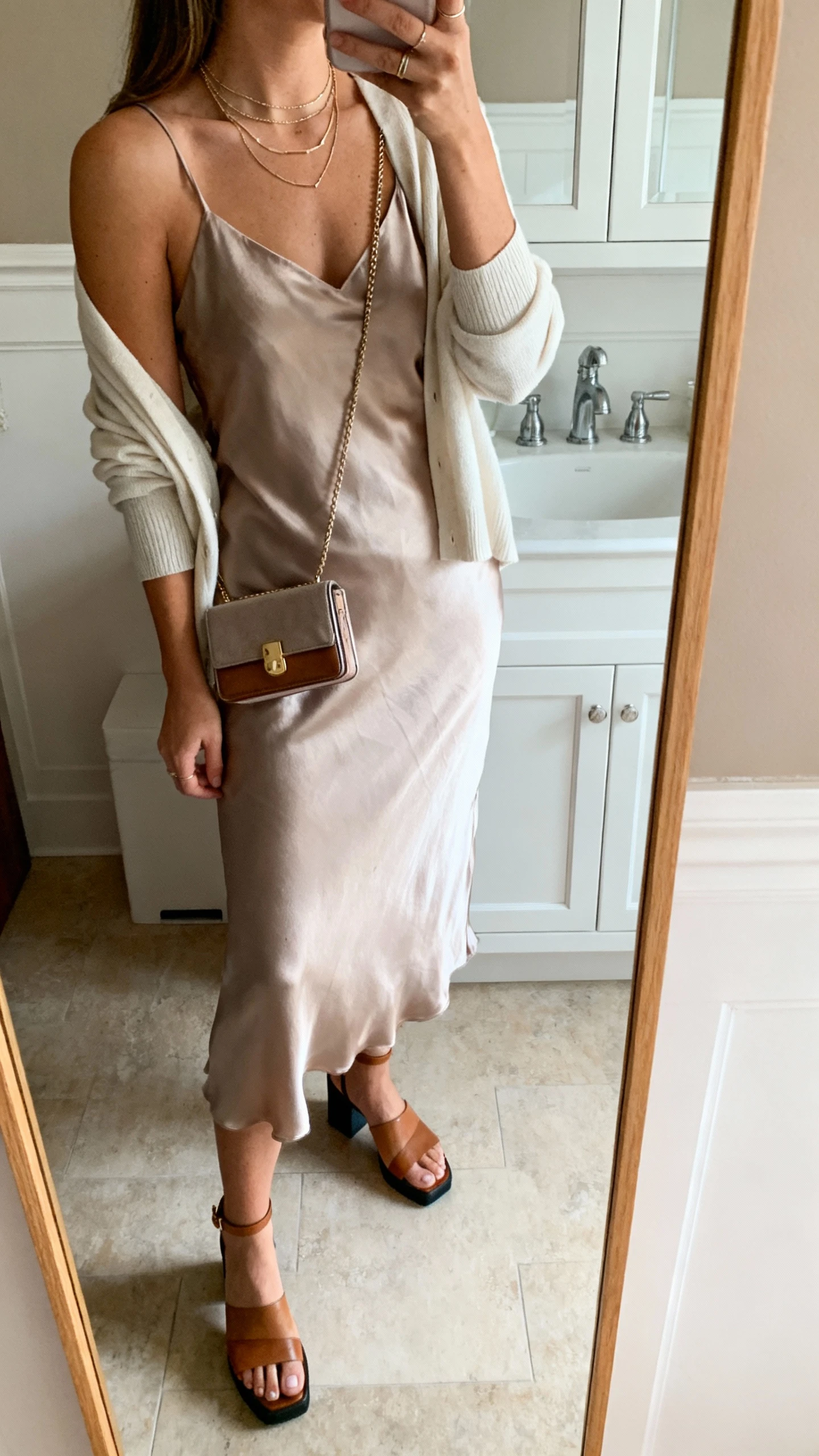 iPhone side-angle mirror selfie of a woman wearing a silky slip dress layered with a light cardigan over the shoulders, low block-heel sandals, a compact crossbody bag, and dainty layered necklaces, face not visible, bathroom mirror, soft indoor lighting, iPhone photo quality.