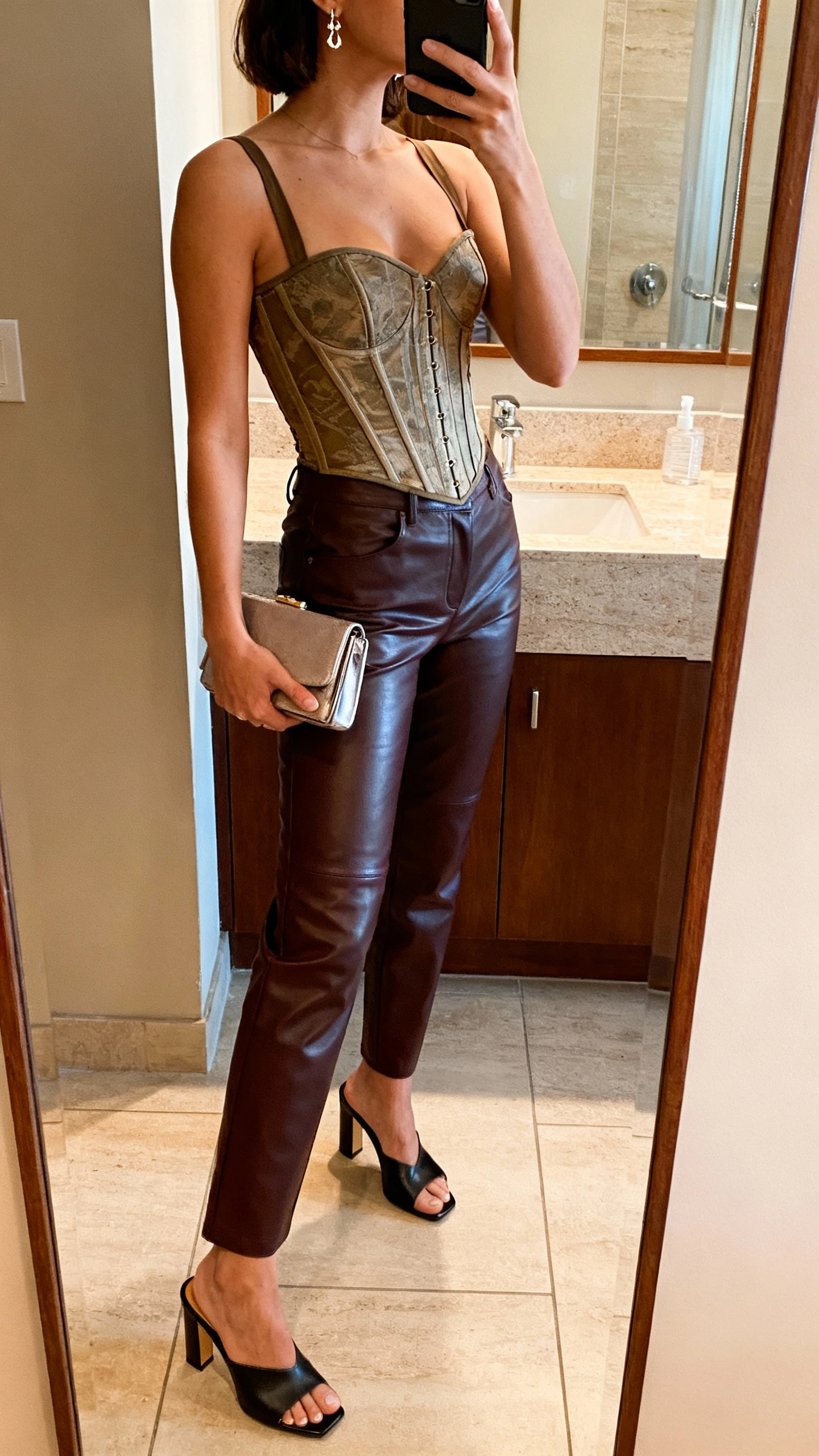 iPhone side-angle mirror selfie of a woman wearing a corset top, leather pants, and heeled mules, small clutch and delicate earrings, face not visible, bathroom mirror with warm indoor lighting, iPhone photo quality.
