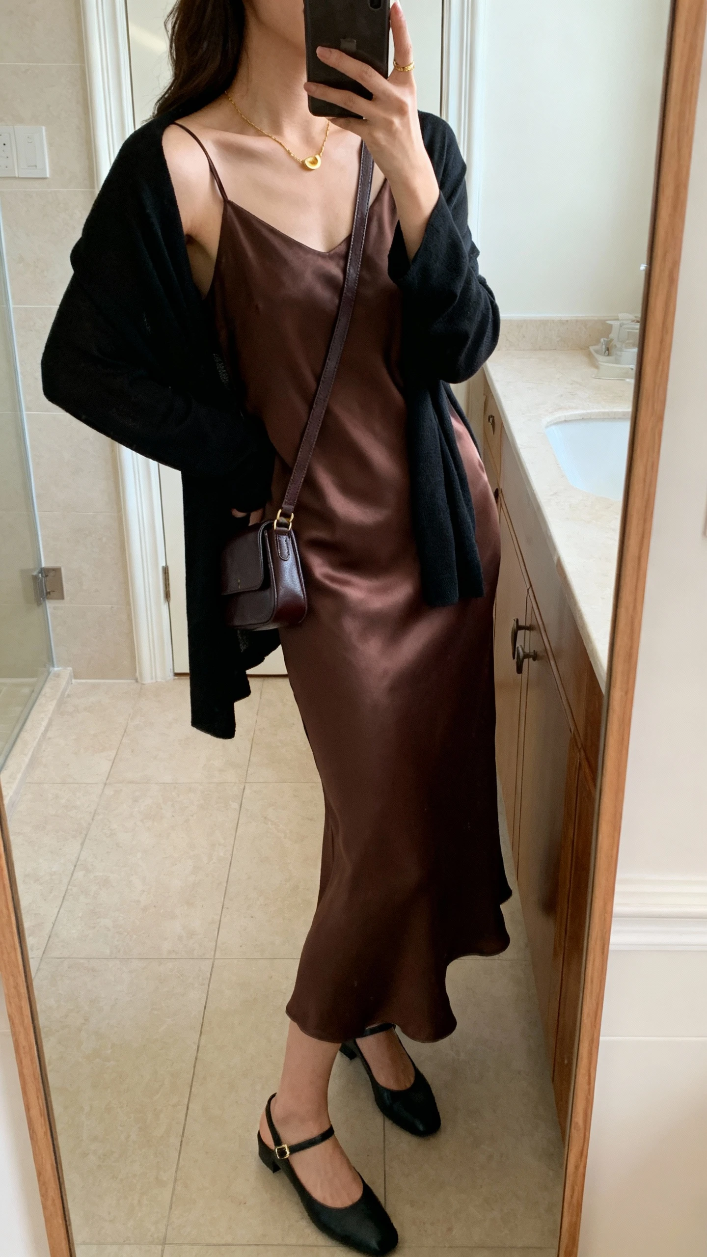 iPhone side-angle mirror selfie of a woman wearing an espresso-brown silk slip dress with a lightweight black cardigan draped over shoulders, delicate gold necklace, black slingback flats, and a small leather crossbody, face not visible, bathroom mirror, soft morning light, iPhone photo quality