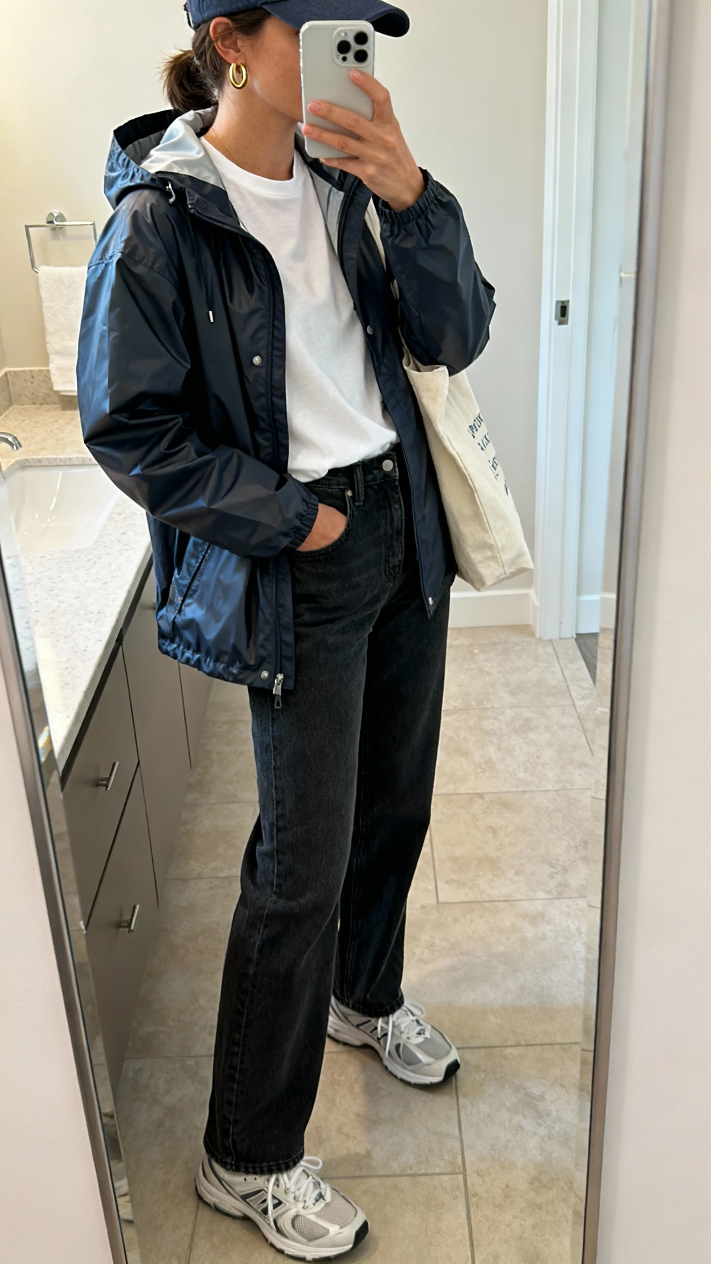 iPhone side-angle mirror selfie of a woman wearing a sporty waterproof rain jacket with a hood, crisp white tee, straight-leg dark jeans, clean sneakers, baseball cap and simple gold hoops, tote bag, face not visible, bathroom mirror, indoor soft light, iPhone photo quality.