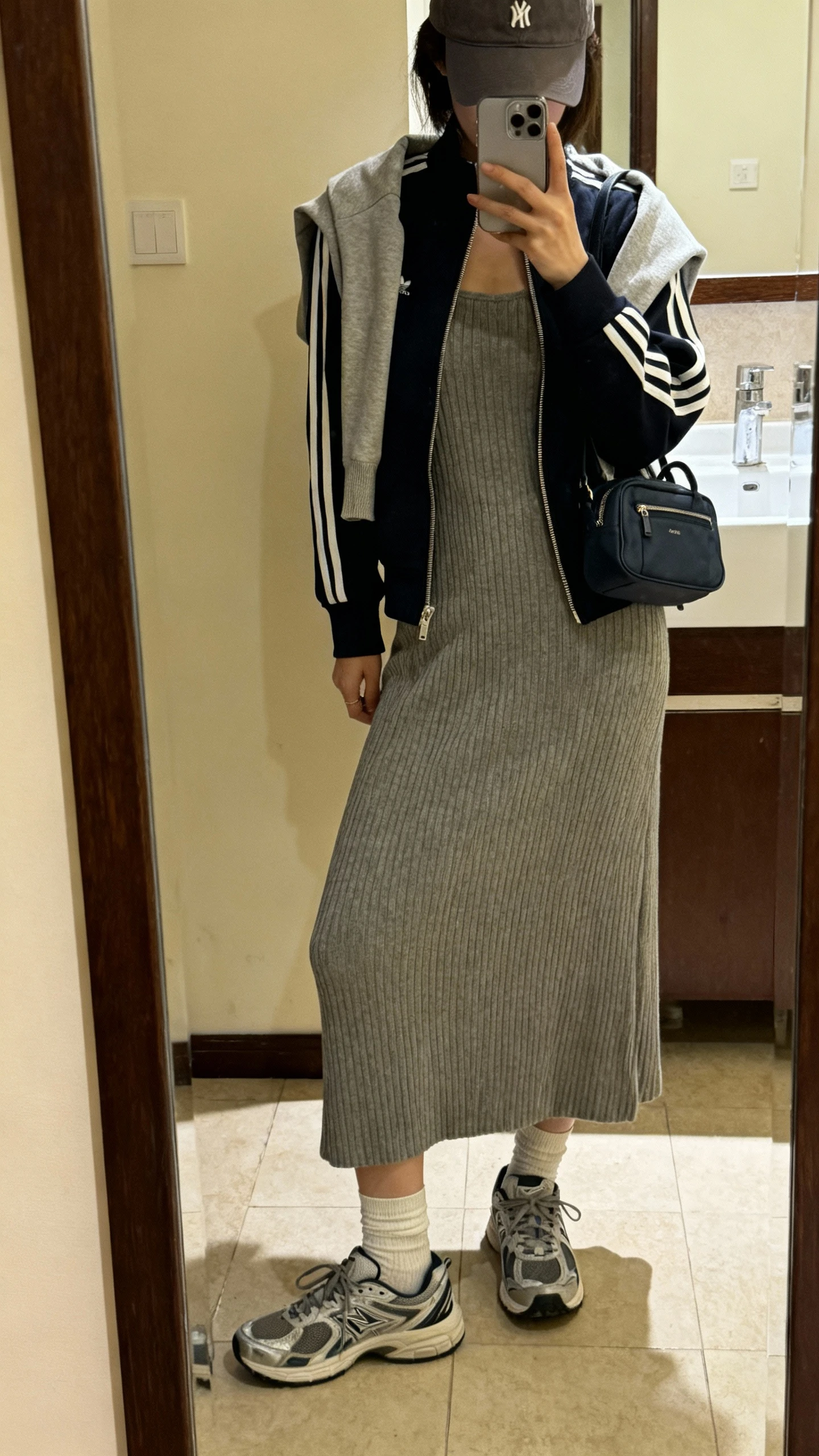 iPhone side-angle mirror selfie of a woman wearing a ribbed knit midi dress with a sporty zip-up track jacket layered over her shoulders, baseball cap, mini backpack, ankle socks, and retro running sneakers; face not visible, bathroom full-length mirror, soft indoor lighting, iPhone photo quality.