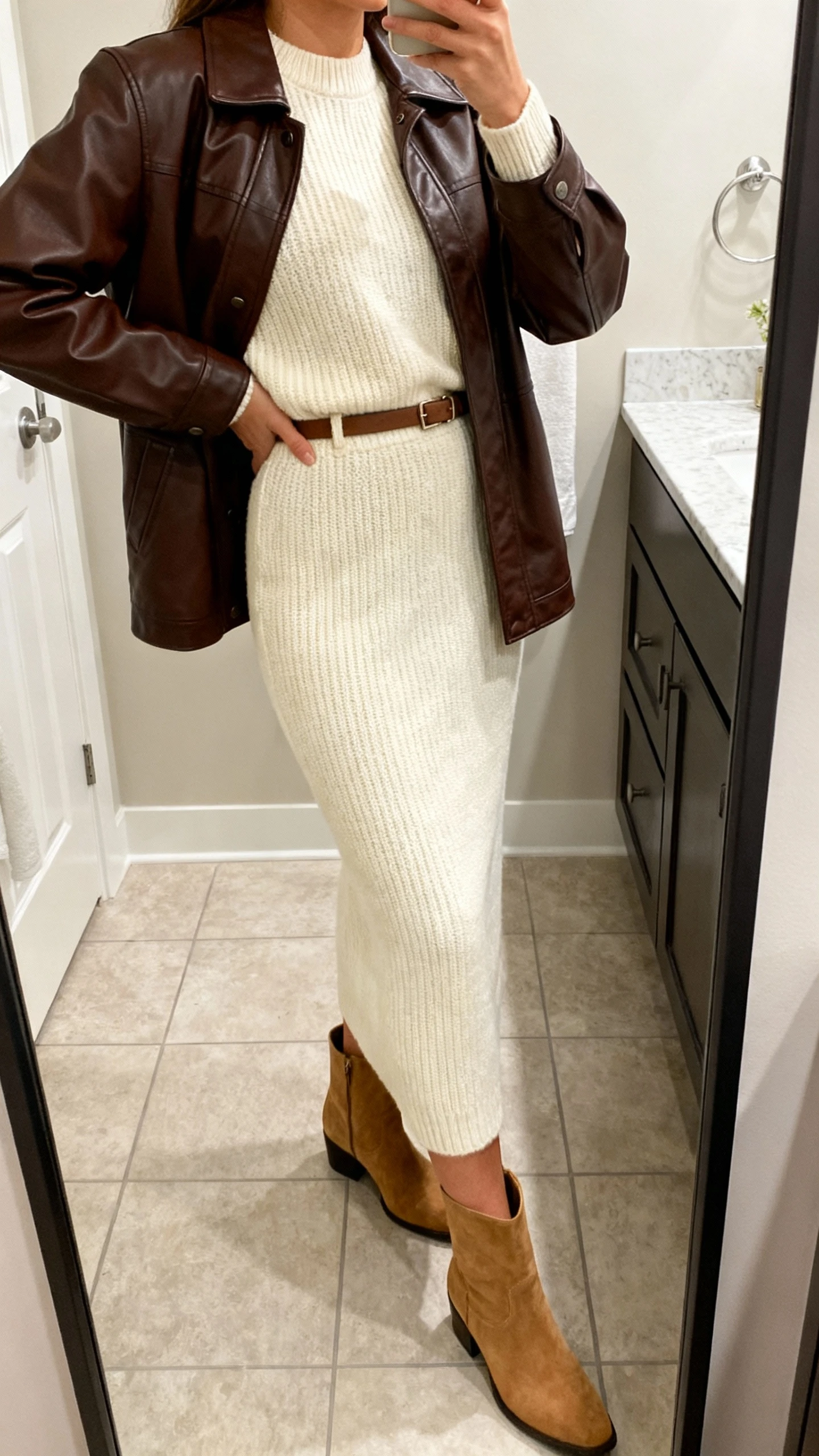 iPhone side-angle mirror selfie of a woman wearing a monochrome cream knit set (crewneck sweater and matching knit skirt) layered with a chocolate brown leather jacket, tan ankle boots, and a slim belt, face not visible, bathroom full-length mirror, soft indoor lighting, iPhone photo quality.