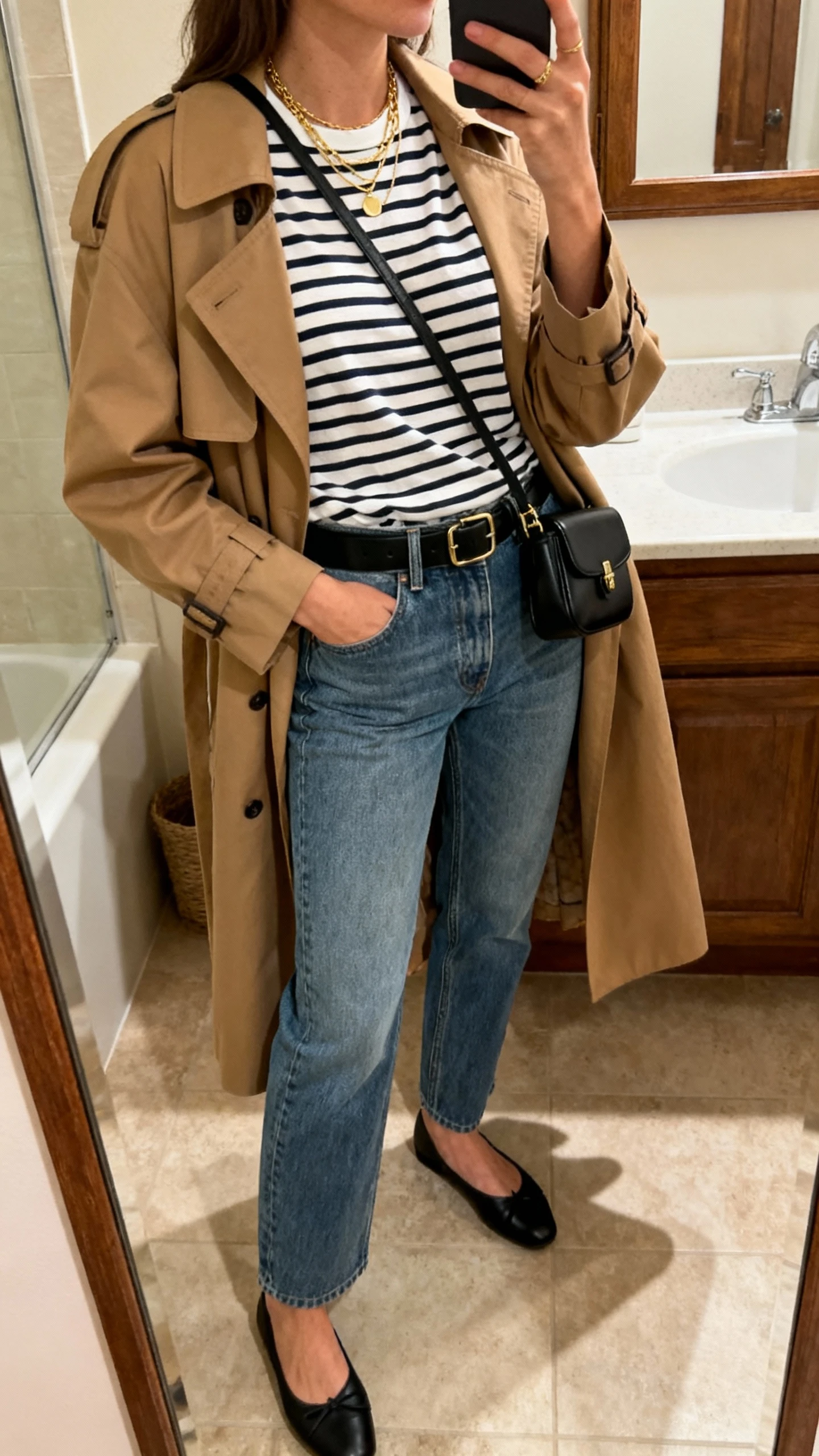 iPhone side-angle mirror selfie of a woman wearing French-girl denim with a celeb twist: straight-leg medium-wash jeans, striped Breton tee tucked in, camel trench coat draped, black ballet flats, black leather belt, small black crossbody, delicate gold layered necklaces, face not visible, bathroom mirror, natural indoor lighting, iPhone photo quality