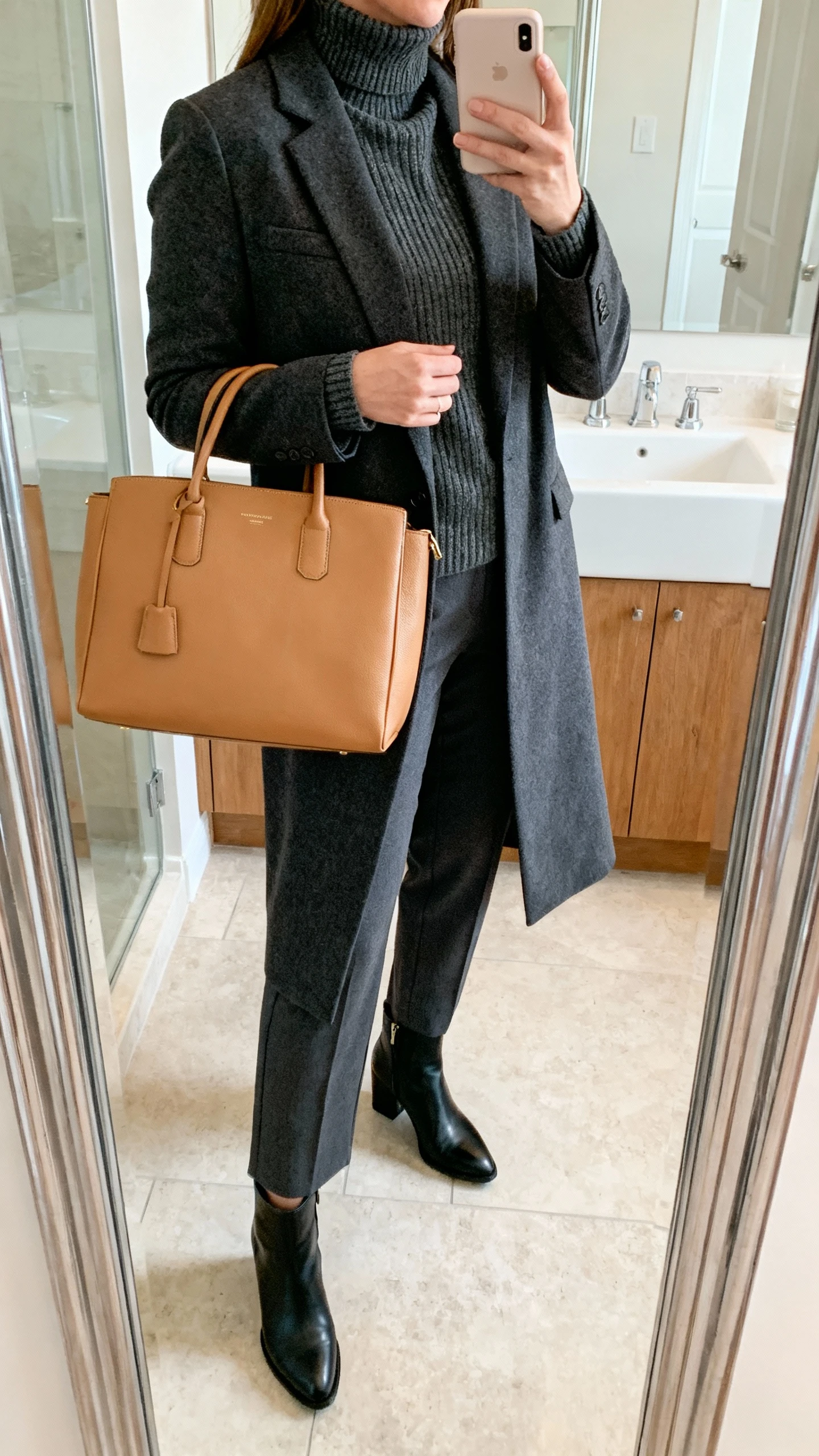 iPhone side-angle mirror selfie of a woman wearing a structured tan tote, monochrome charcoal knit turtleneck, matching charcoal longline blazer, charcoal straight-leg trousers, and sleek black ankle boots; face not visible, bathroom mirror, soft morning light, iPhone photo quality.
