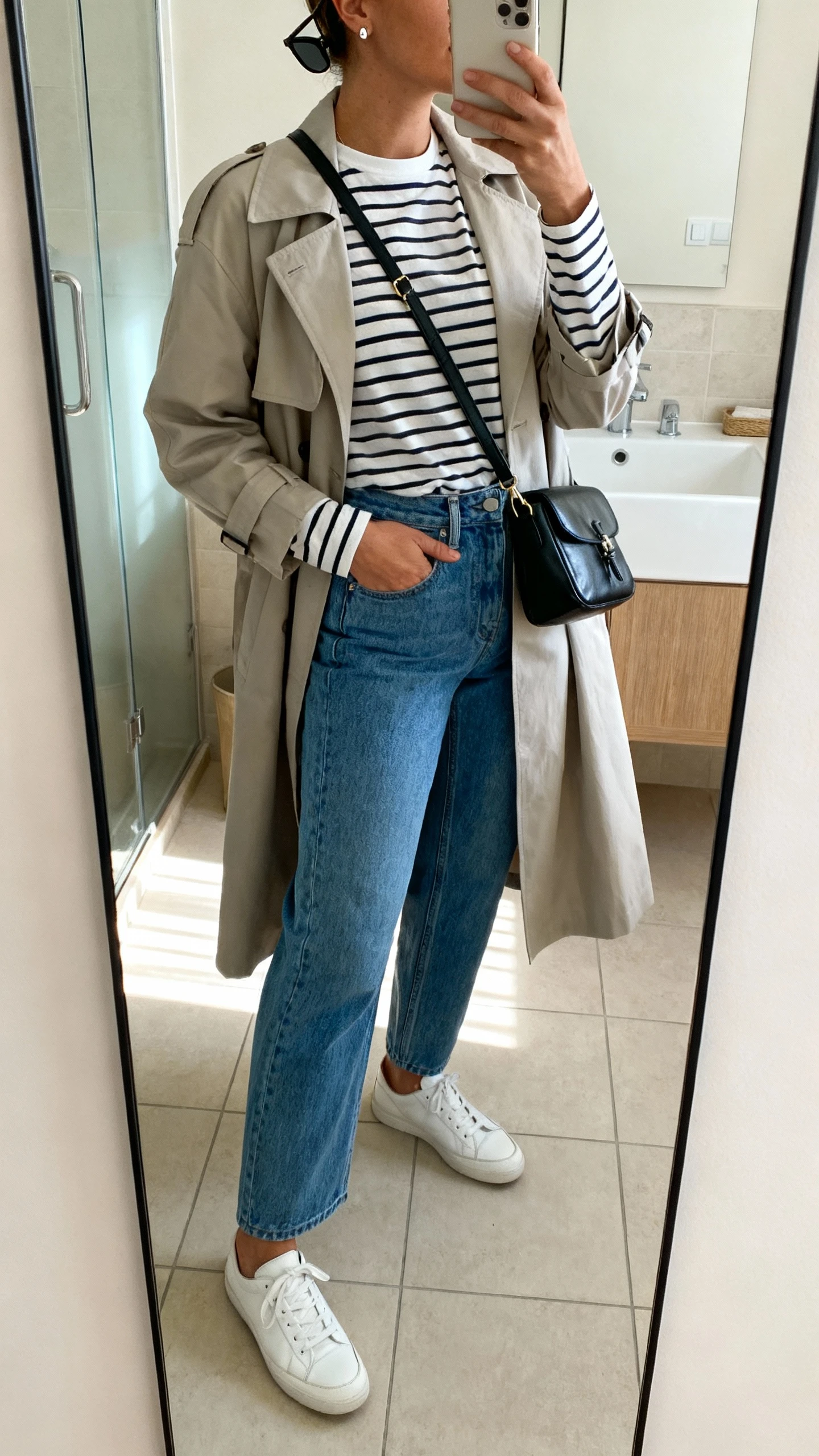iPhone side-angle mirror selfie of a woman wearing a striped long-sleeve tee, high-waisted straight-leg blue jeans, lightweight trench coat, white leather sneakers, black leather crossbody bag worn across chest, simple stud earrings and sunglasses on head, face not visible, bathroom mirror, soft morning light, iPhone photo quality.