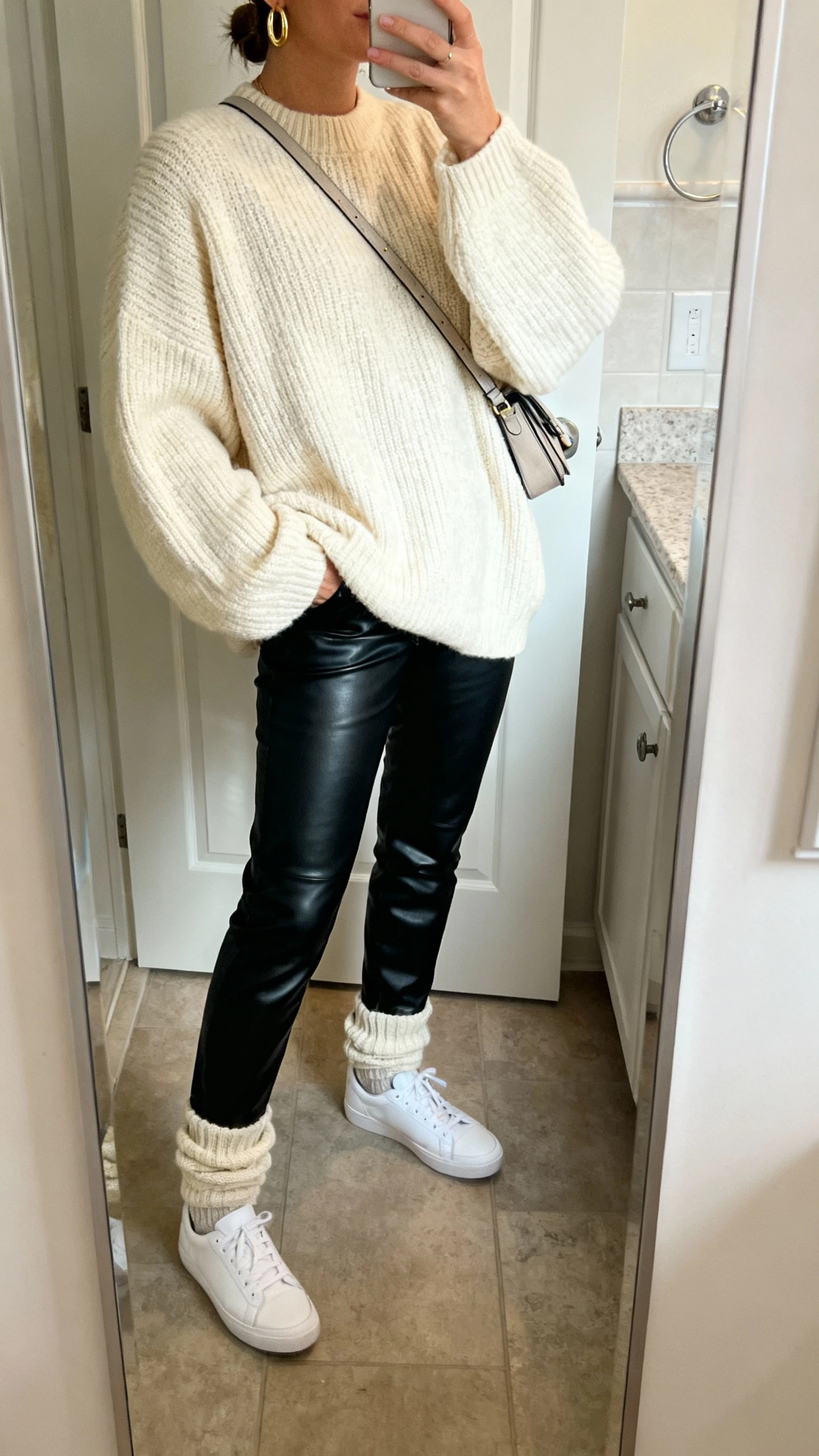 iPhone side-angle mirror selfie of a woman wearing black leather pants, a cozy oversized cream knit sweater, white low-top sneakers, slouchy socks, crossbody bag, and simple gold hoops, face not visible, bathroom full-length mirror, soft morning light, iPhone photo quality.