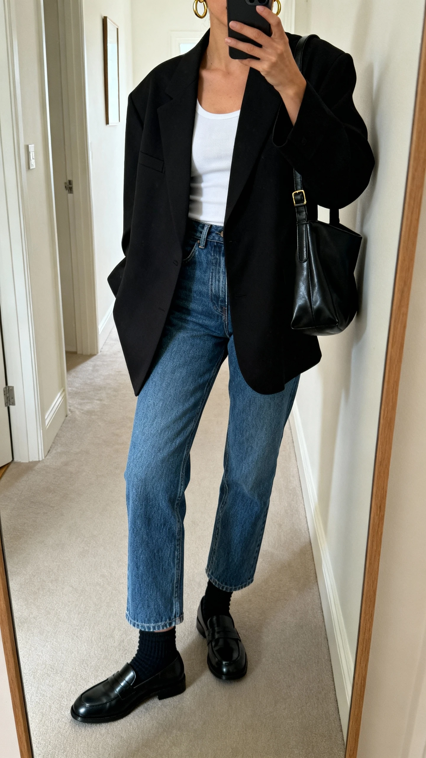 iPhone side-angle mirror selfie of a woman wearing straight-leg blue jeans, a black oversized blazer over a white fitted tank, black loafers with socks, minimalist gold hoops, and a black leather shoulder bag; face not visible, cropped at shoulders, full-length hallway mirror, soft indoor lighting, iPhone photo quality.