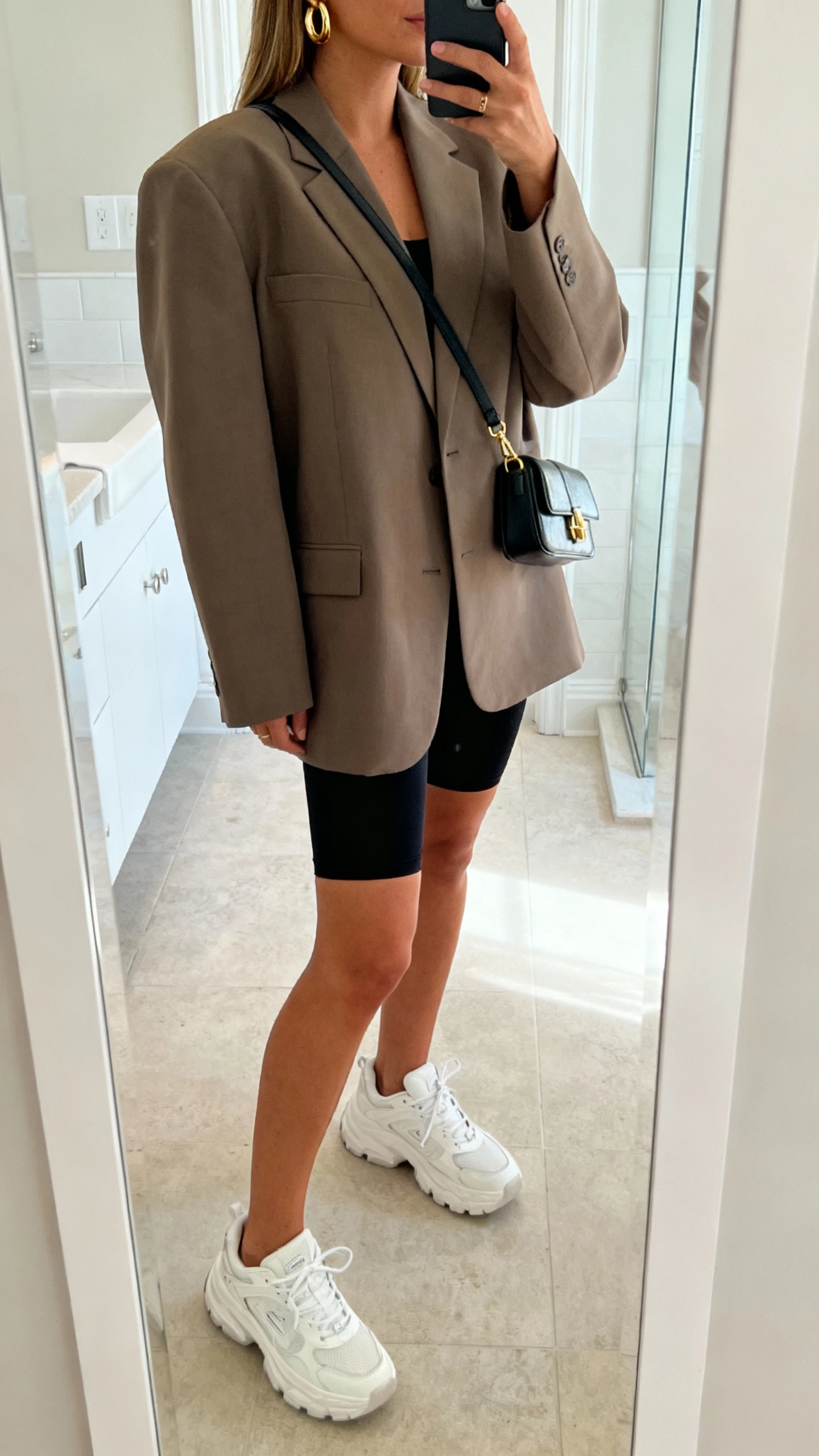 iPhone side-angle mirror selfie of a woman wearing an oversized boxy blazer, black bike shorts, chunky white dad sneakers, gold hoops, and a mini shoulder bag, face not visible, cropped at shoulders, bathroom full-length mirror with bright indoor lighting, iPhone photo quality.