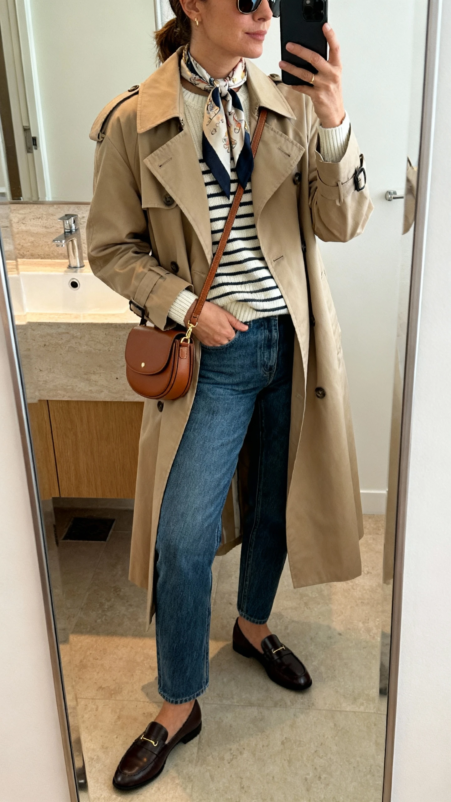 iPhone side-angle mirror selfie of a woman wearing a classic trench coat over a lightweight striped knit, straight-leg jeans, loafers, silk neck scarf, crossbody saddle bag, and slim sunglasses, face not visible, bathroom mirror with soft indoor lighting, iPhone photo quality