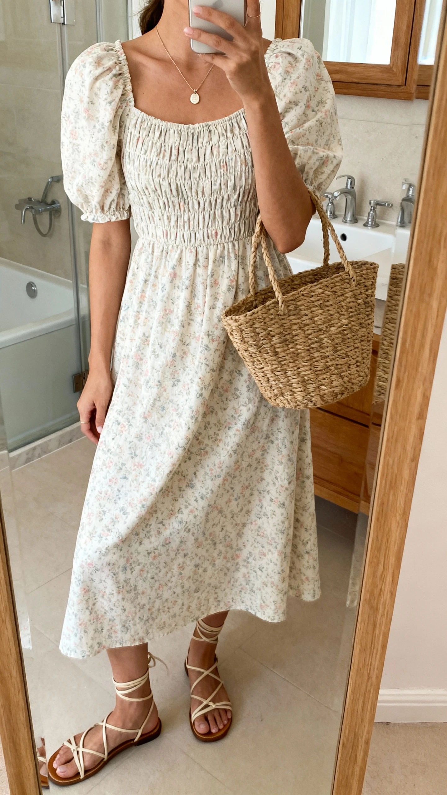 iPhone side-angle mirror selfie of a woman wearing a romantic organic cotton dress: midi-length puff-sleeve dress with smocked bodice, soft floral print, strappy sandals, dainty pendant necklace, woven basket bag; face not visible, bathroom mirror with soft indoor lighting, iPhone photo quality.