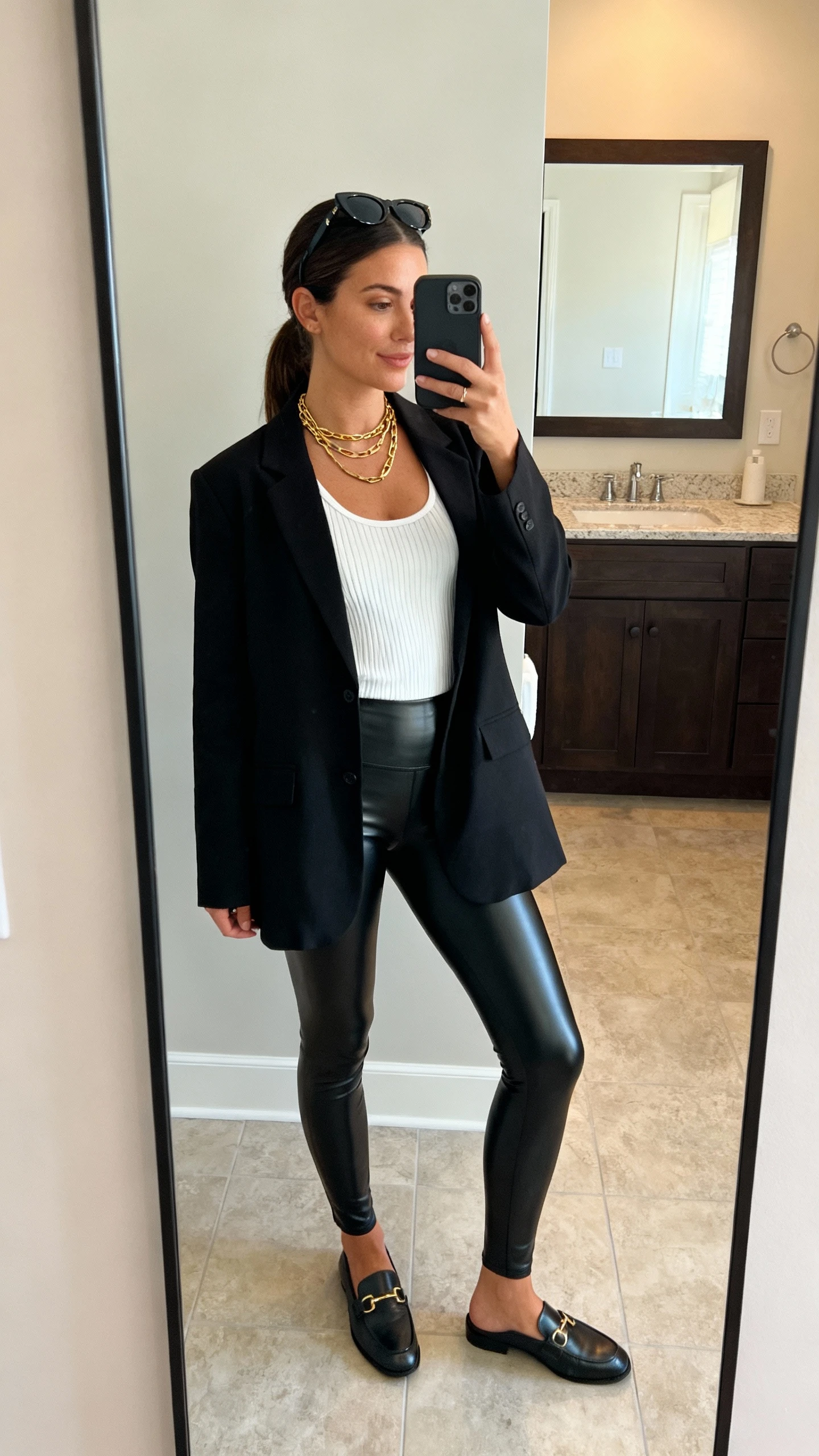 iPhone side-angle mirror selfie of a woman wearing faux leather leggings, a relaxed-fit black blazer over a white ribbed tank, sleek sunglasses pushed to head, layered gold necklace, and loafers, face not visible, bathroom full-length mirror with soft indoor lighting, iPhone photo quality.