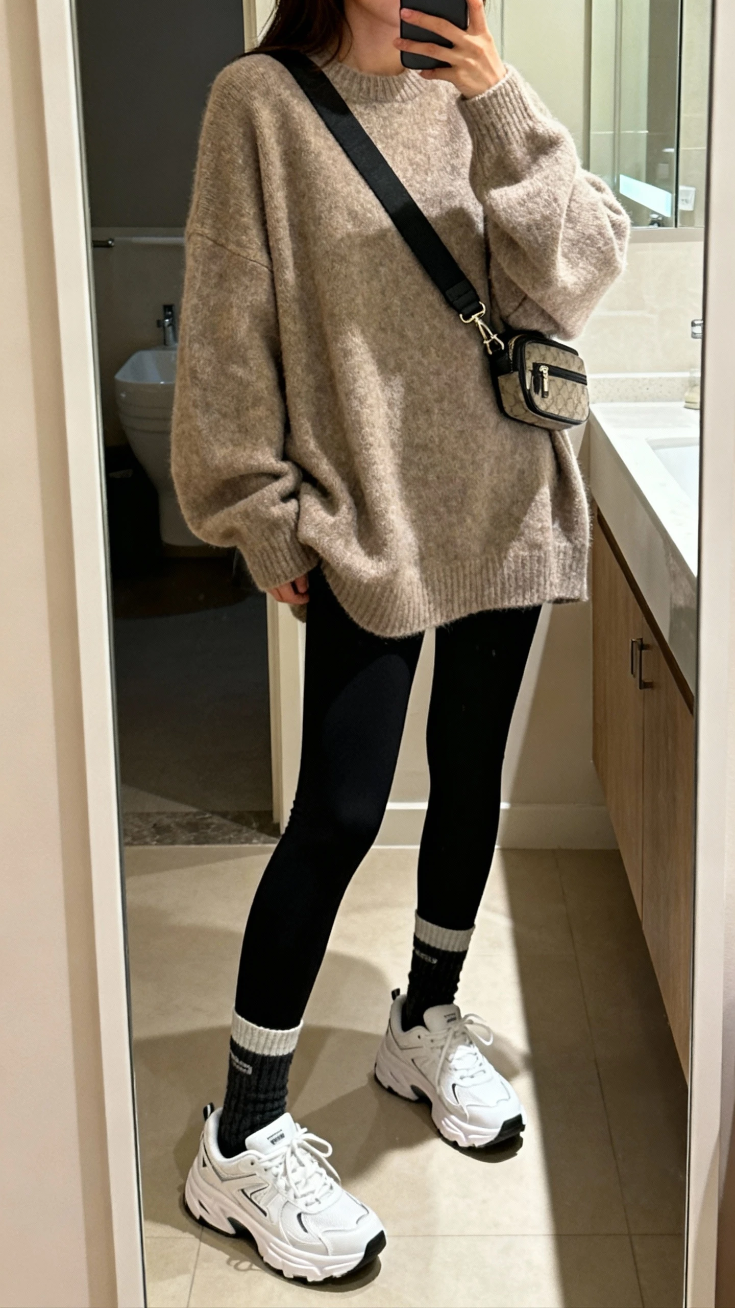 iPhone side-angle mirror selfie of a woman wearing an oversized slouchy sweater, black leggings, crew socks, white dad sneakers, and a crossbody belt bag, face not visible, cropped at shoulders, bathroom mirror with soft indoor lighting, iPhone photo quality.
