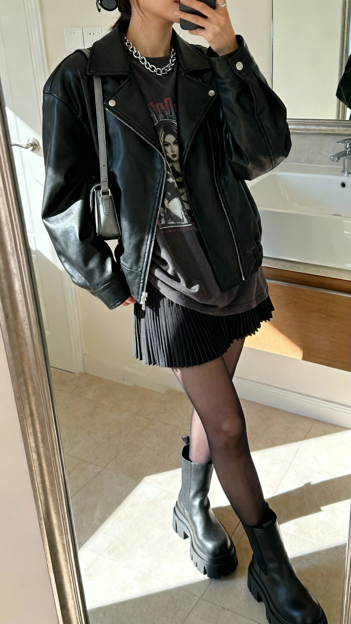 iPhone side-angle mirror selfie of a woman wearing a black leather jacket over a slouchy graphic tee, micro-pleated mini skirt, sheer black tights, chunky platform boots, silver chain necklace, smudgy eyeliner-look sunglasses perched on head, and a small shoulder bag, face not visible, bathroom mirror, indoor morning light, iPhone photo quality.