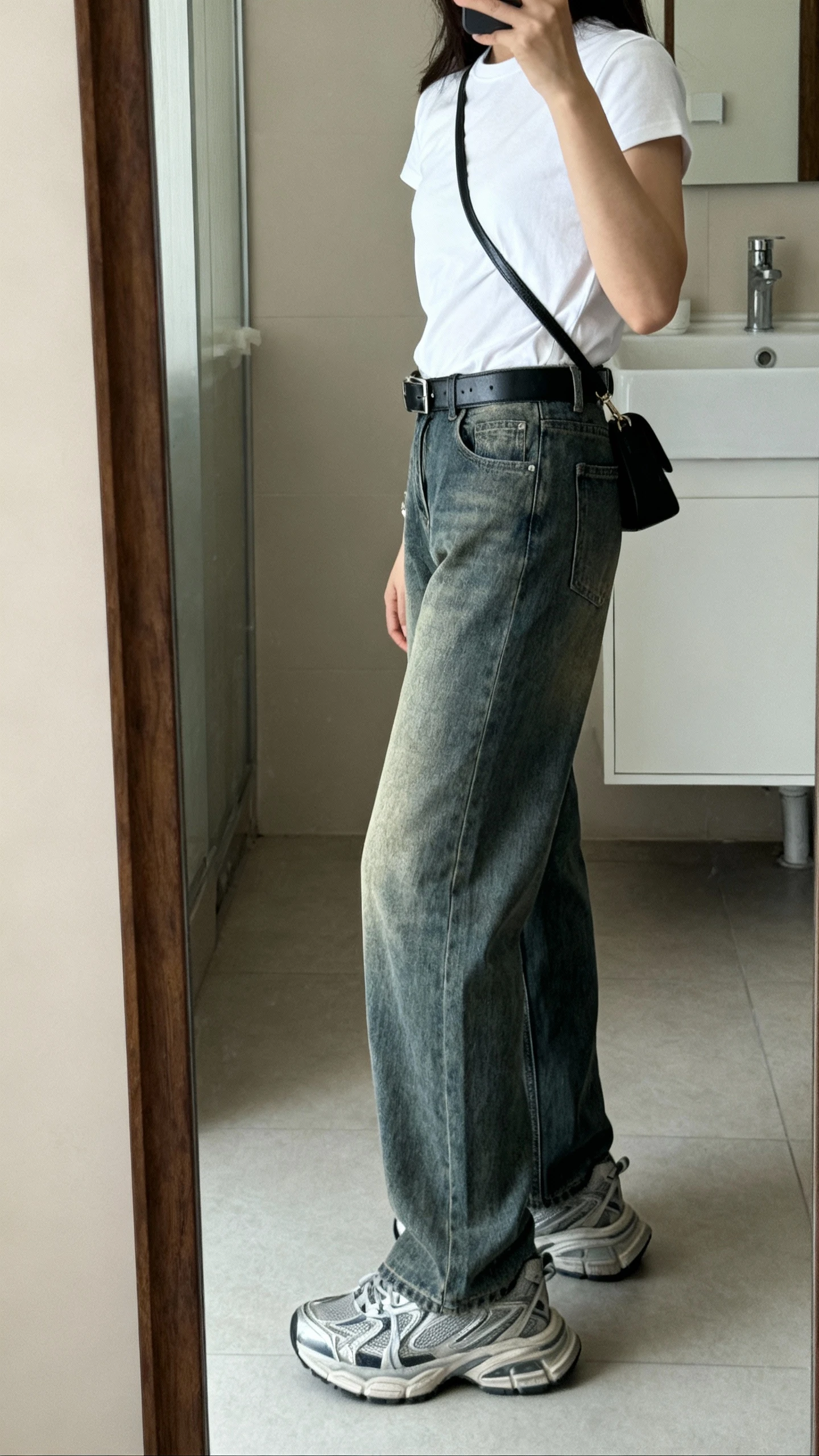 iPhone side-angle mirror selfie of a woman wearing straight-leg vintage-wash denim, tucked-in plain white tee, chunky dad sneakers, black leather belt, simple crossbody bag, face not visible, bathroom full-length mirror with soft indoor lighting, iPhone photo quality.