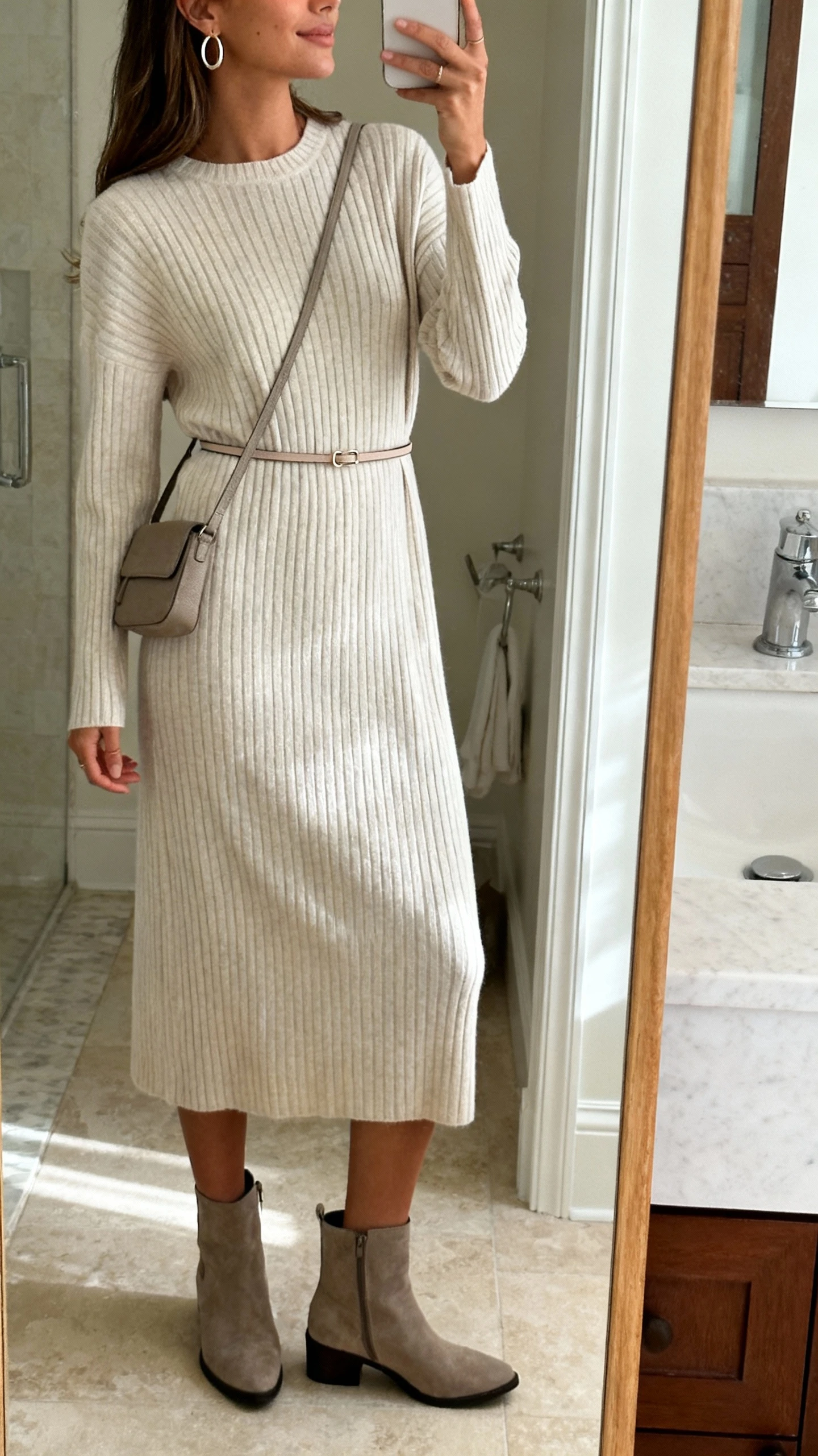 iPhone side-angle mirror selfie of a woman wearing an effortless ribbed knit midi dress, thin waist belt, ankle boots, simple hoop earrings, and a small crossbody bag, face not visible, bathroom full-length mirror with soft indoor light, iPhone photo quality.