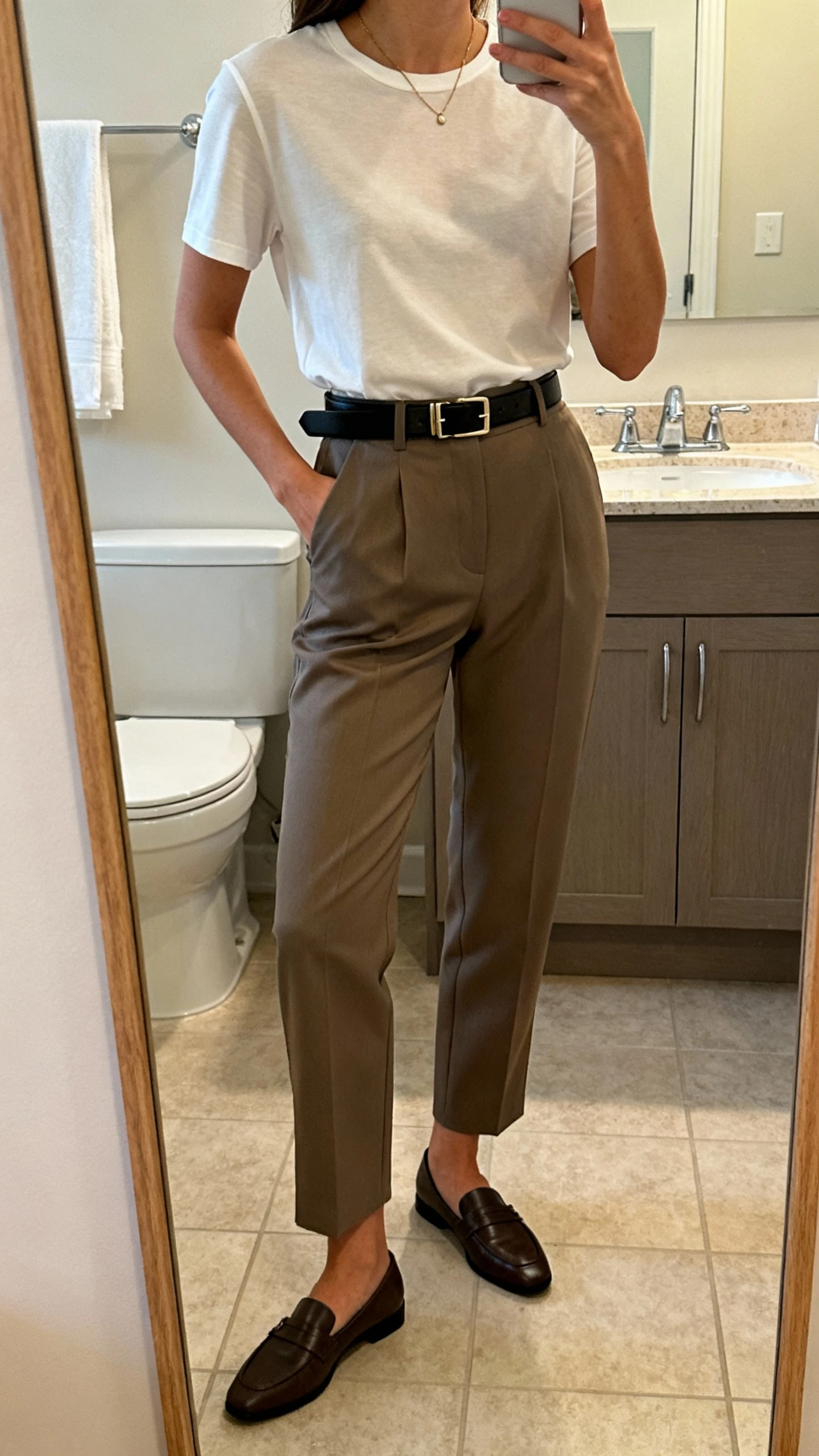 iPhone side-angle mirror selfie of a woman wearing tailored trousers with an elevated tee tucked in, sleek belt, loafers, and a delicate necklace, face not visible, bathroom full-length mirror, soft indoor lighting, iPhone photo quality.