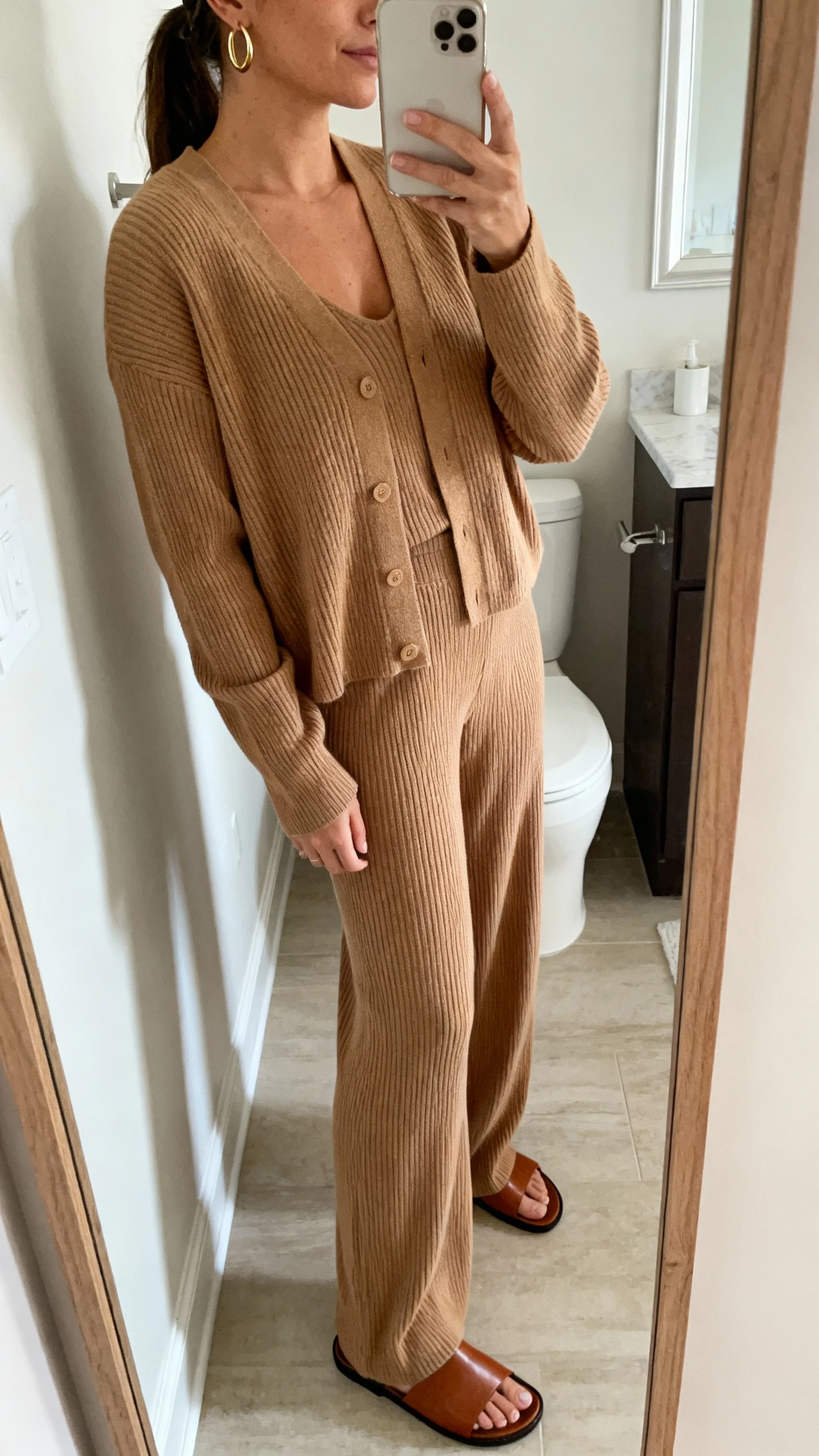 iPhone side-angle mirror selfie of a woman wearing a matching ribbed knit set: camel cardigan and coordinating wide-leg knit pants with minimalist gold hoops and sleek leather slides, face not visible, bathroom full-length mirror, soft morning light, iPhone photo quality.