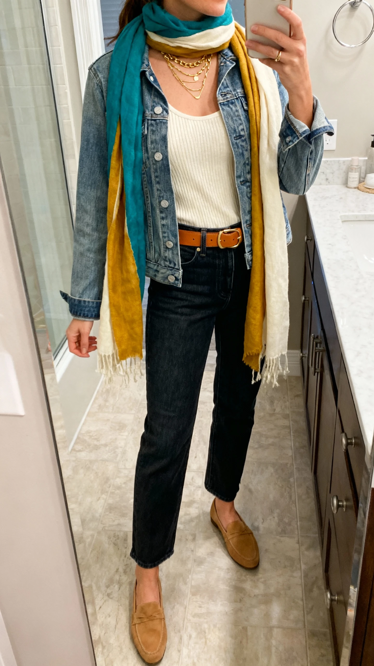 iPhone side-angle mirror selfie of a woman wearing a medium-wash denim jacket, cream knit tank, high-waisted dark denim jeans, tan loafers, a color-harmony scarf in teal, mustard, and cream draped loosely, gold layered necklaces, and a tan belt, face not visible, bathroom full-length mirror, morning indoor lighting, iPhone photo quality.