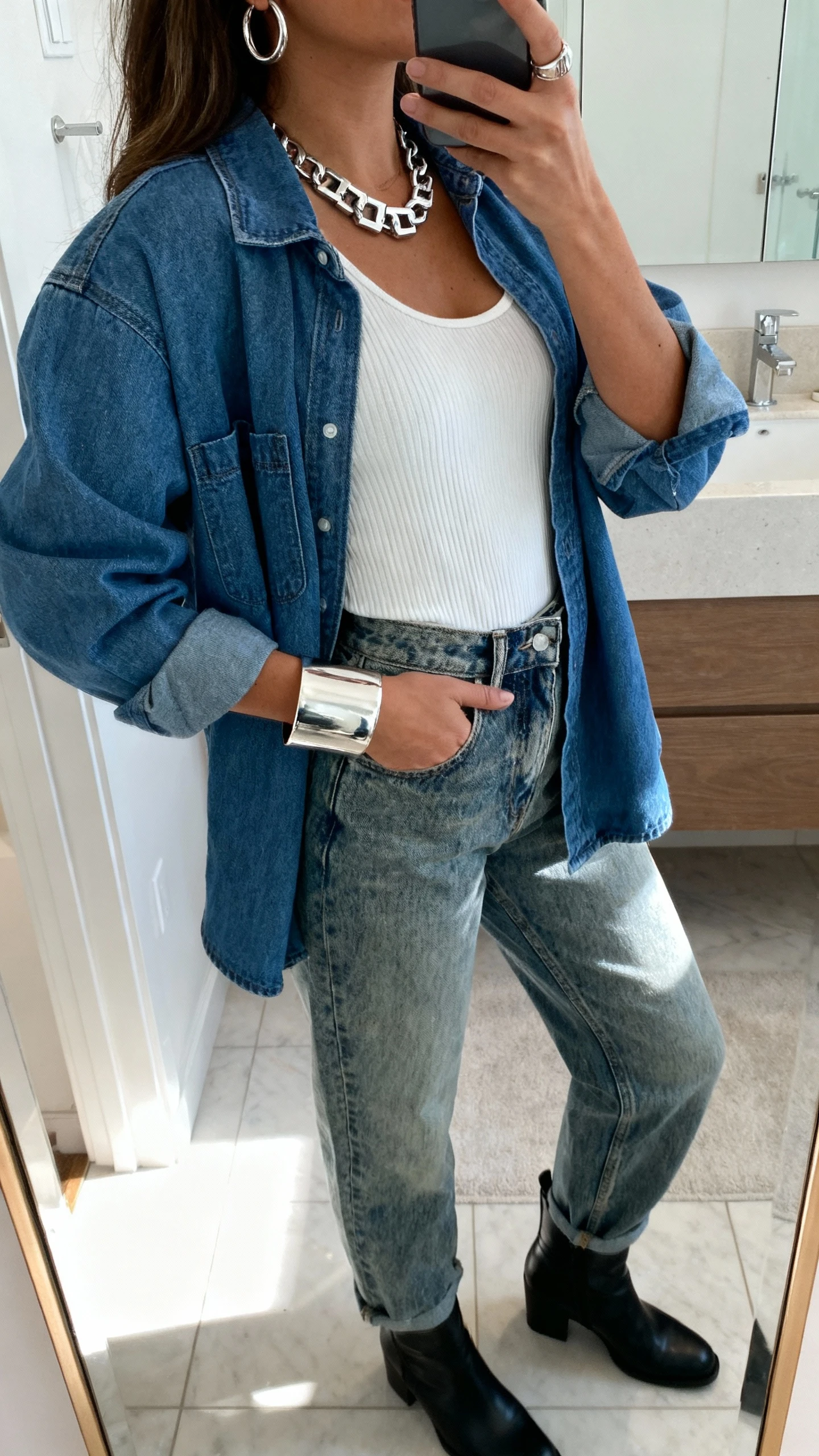 iPhone side-angle mirror selfie of a woman wearing a relaxed blue denim jacket, white ribbed tank, high-waist vintage wash jeans, chunky silver jewelry (wide cuff bracelet, thick huggie hoops, box chain), and black ankle boots, face not visible, bathroom mirror, morning light, iPhone photo quality