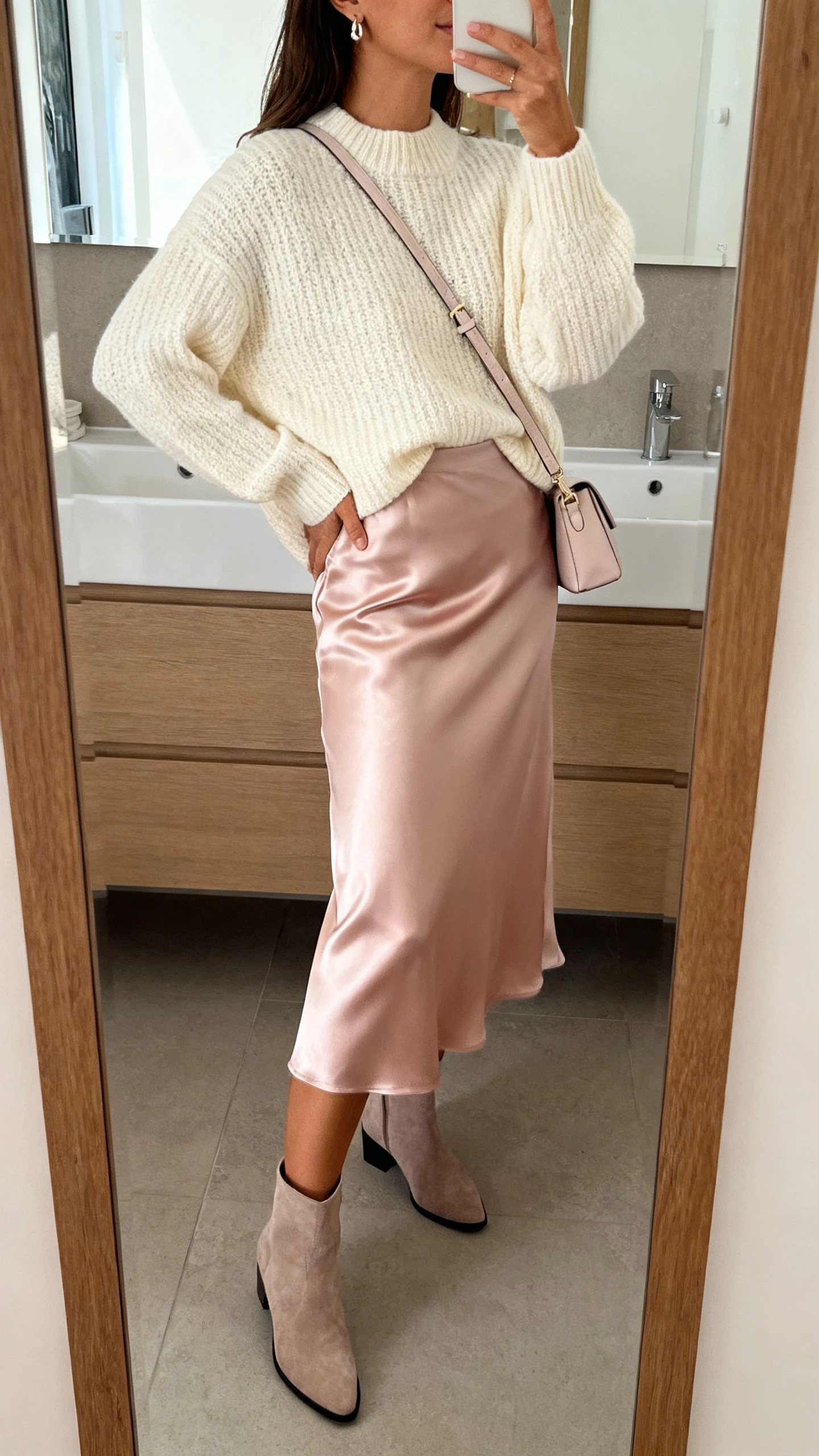 iPhone side-angle mirror selfie of a woman wearing a cozy cream knit sweater tucked into a blush satin midi skirt, ankle boots, dainty earrings, and a soft crossbody bag, face not visible, bathroom mirror, morning indoor light, iPhone photo quality.