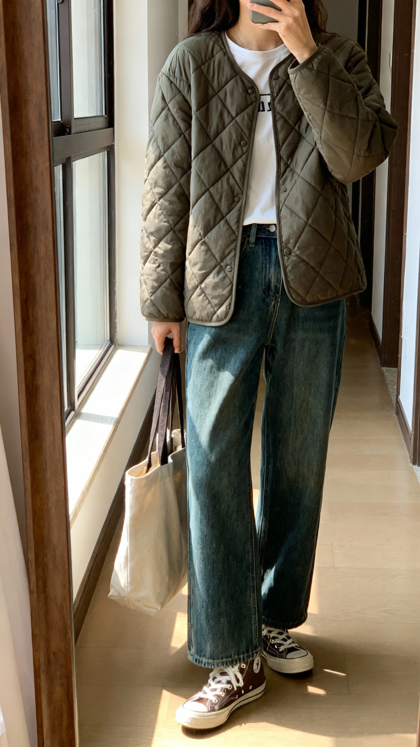 iPhone side-angle mirror selfie of a woman wearing a quilted jacket over a simple tee with wide-leg jeans and retro sneakers, tote bag tucked under arm, face not visible, hallway full-length mirror, morning window light, iPhone photo quality.