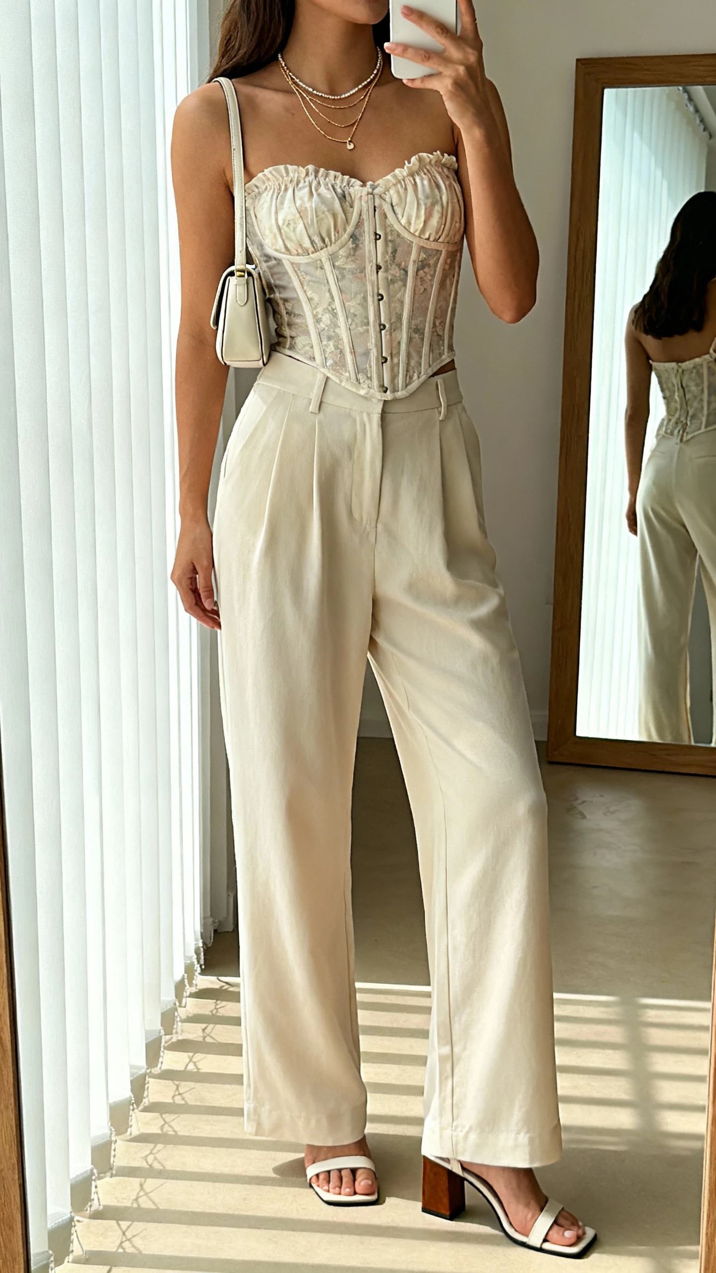iPhone side-angle mirror selfie of a woman wearing a romantic corset top with wide-leg trousers, dainty layered necklaces, mini shoulder bag, and block-heel sandals; face not visible, cropped at shoulders, full-length mirror, morning window light, iPhone photo quality.