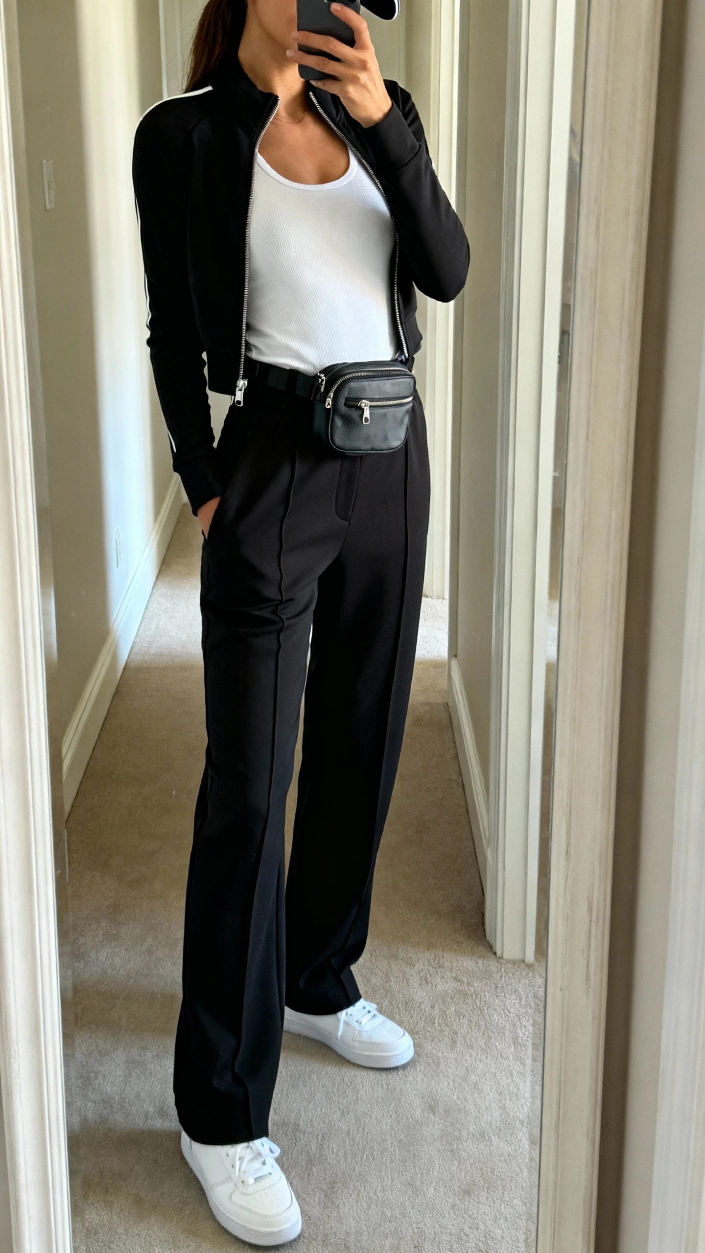 iPhone side-angle mirror selfie of a woman wearing elevated athleisure: black tailored joggers, fitted zip-up track jacket over a white tank, clean white sneakers, baseball cap, mini belt bag, face not visible, hallway mirror, natural daylight, iPhone photo quality.