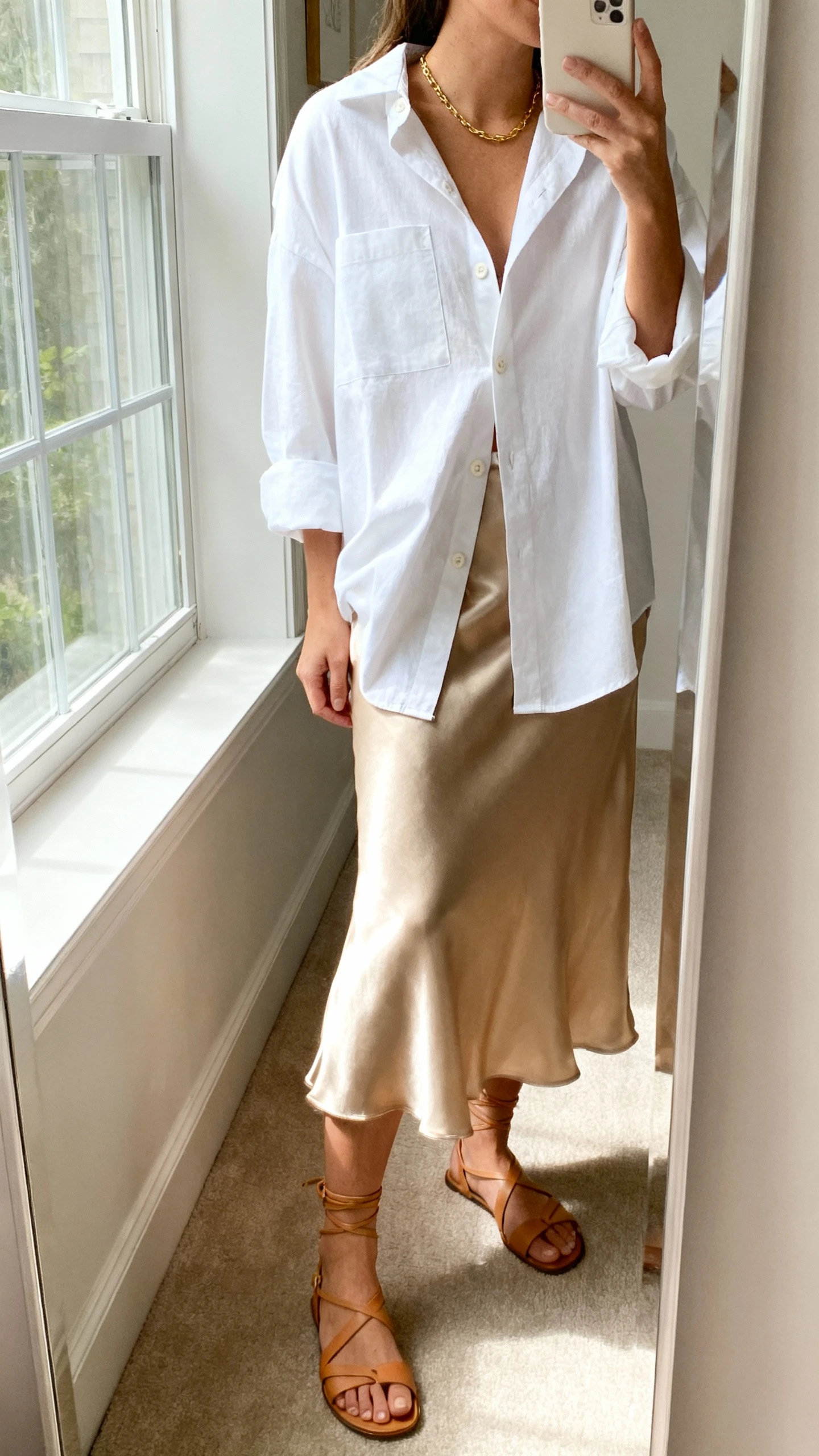 iPhone side-angle mirror selfie of a woman wearing a champagne satin slip skirt with a slightly oversized white boxy button-up half-tucked, paired with strappy tan sandals and a delicate gold necklace, face not visible, cropped at neck, full-length mirror, soft indoor window light, iPhone photo quality.