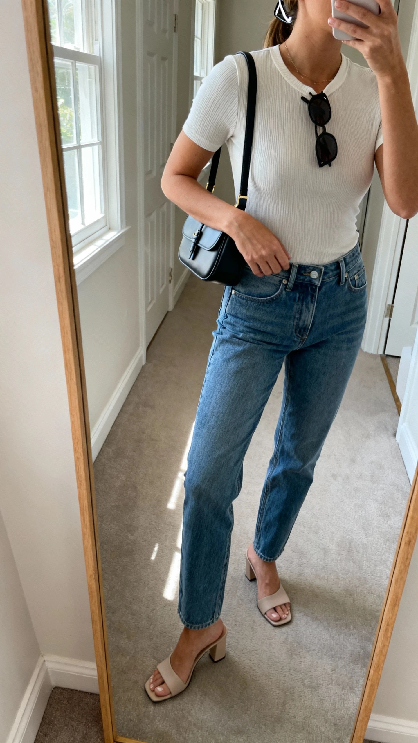 iPhone side-angle mirror selfie of a woman wearing a sleek mini shoulder bag, fitted ribbed baby tee, low-rise straight jeans, slim sunglasses pushed up on head, and minimalist heeled mules; face not visible, cropped at shoulders, hallway mirror, natural window light, iPhone photo quality.