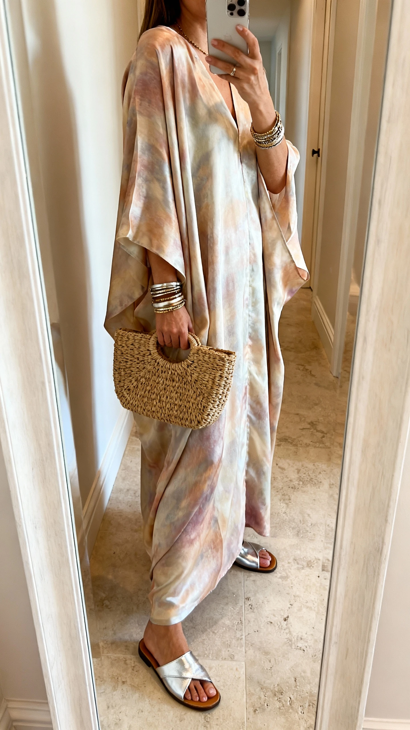 iPhone side-angle mirror selfie of a woman wearing an elevated silk caftan with metallic flat sandals, layered bangles, and a structured raffia clutch, face not visible, cropped at shoulders, hallway mirror with soft indoor lighting, iPhone photo quality.