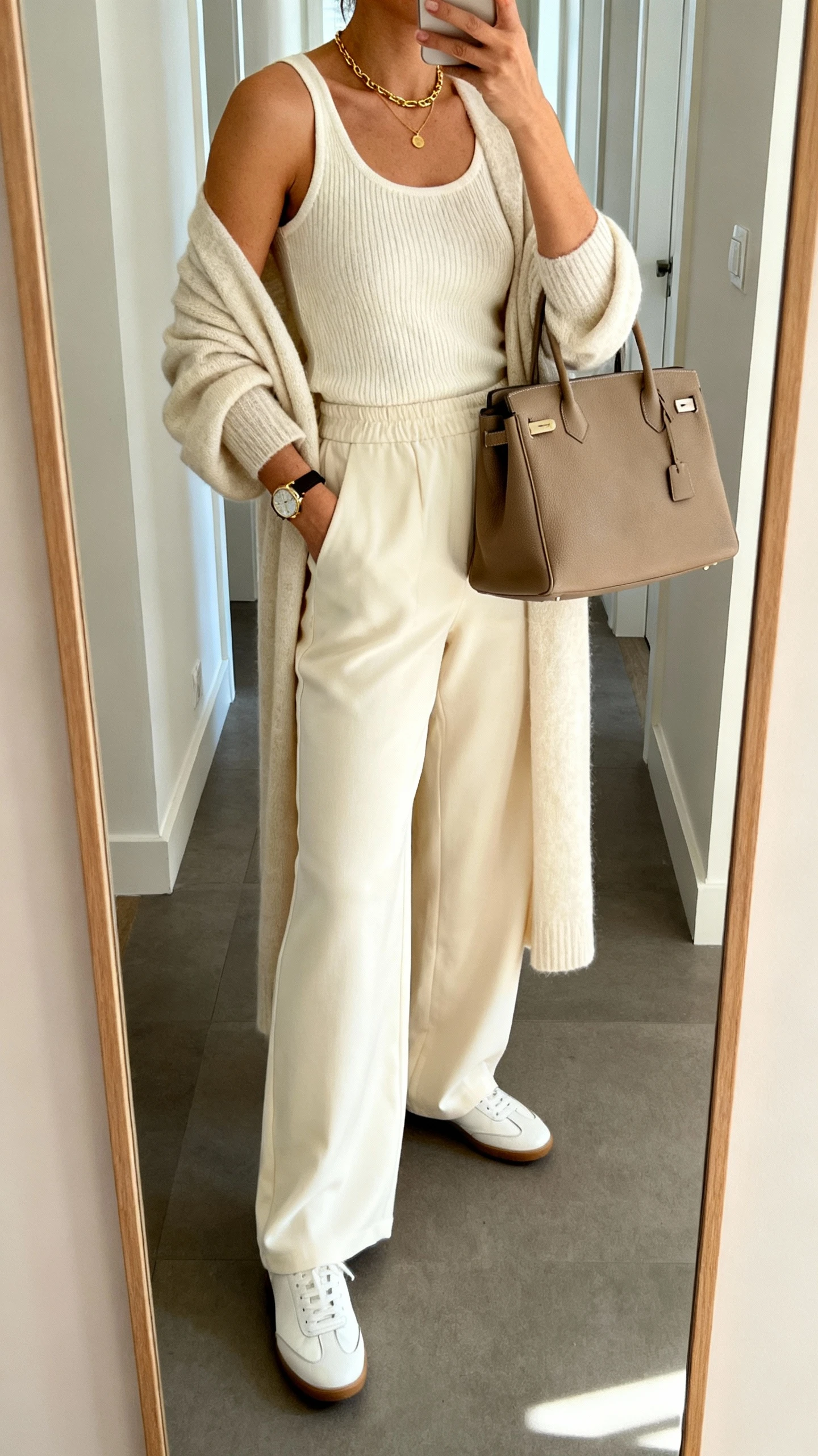 iPhone side-angle mirror selfie of a woman wearing a cream knit tank, matching cream wide-leg trousers, longline matching cardigan, minimalist white leather sneakers, structured taupe tote, delicate gold necklace and watch, face not visible, cropped at shoulders, hallway full-length mirror, soft morning light, iPhone photo quality.