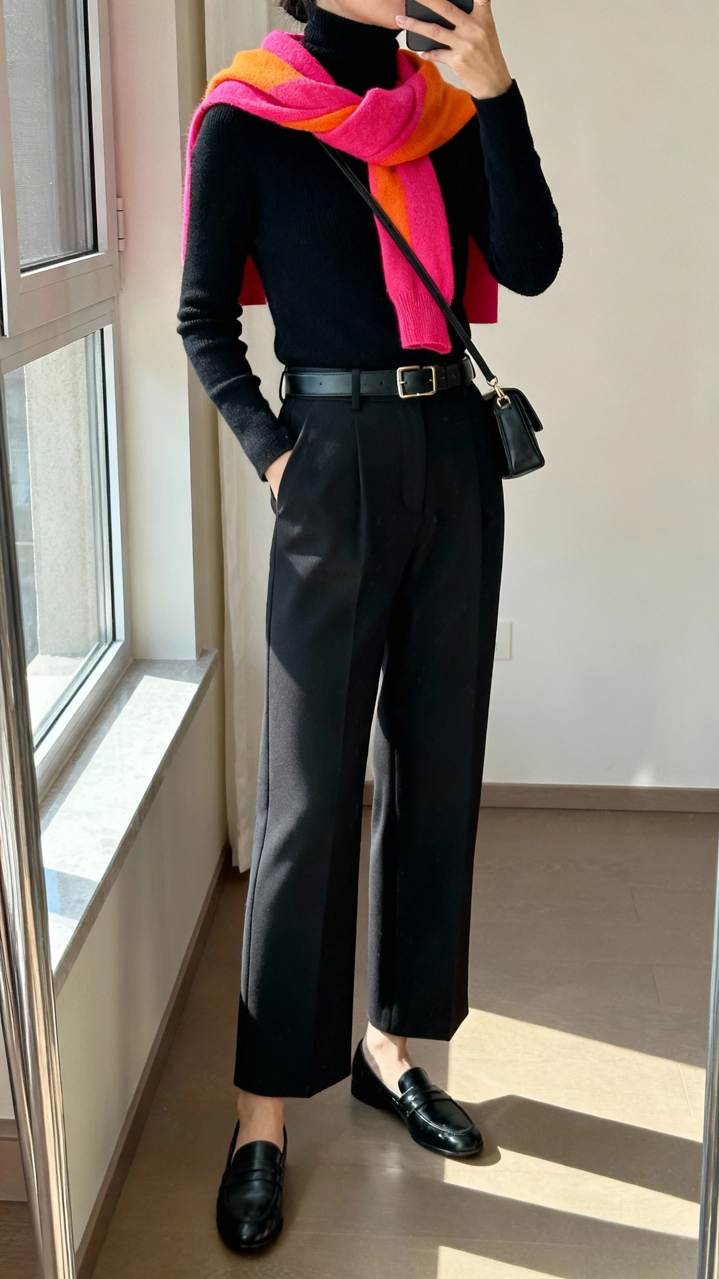 iPhone side-angle mirror selfie of a woman wearing a monochrome black turtleneck, black tailored wide-leg pants, black loafers, sleek belt, and a bright color-pop scarf draped neatly, small crossbody bag, face not visible, cropped at shoulders, full-length mirror by a window, natural light, iPhone photo quality.