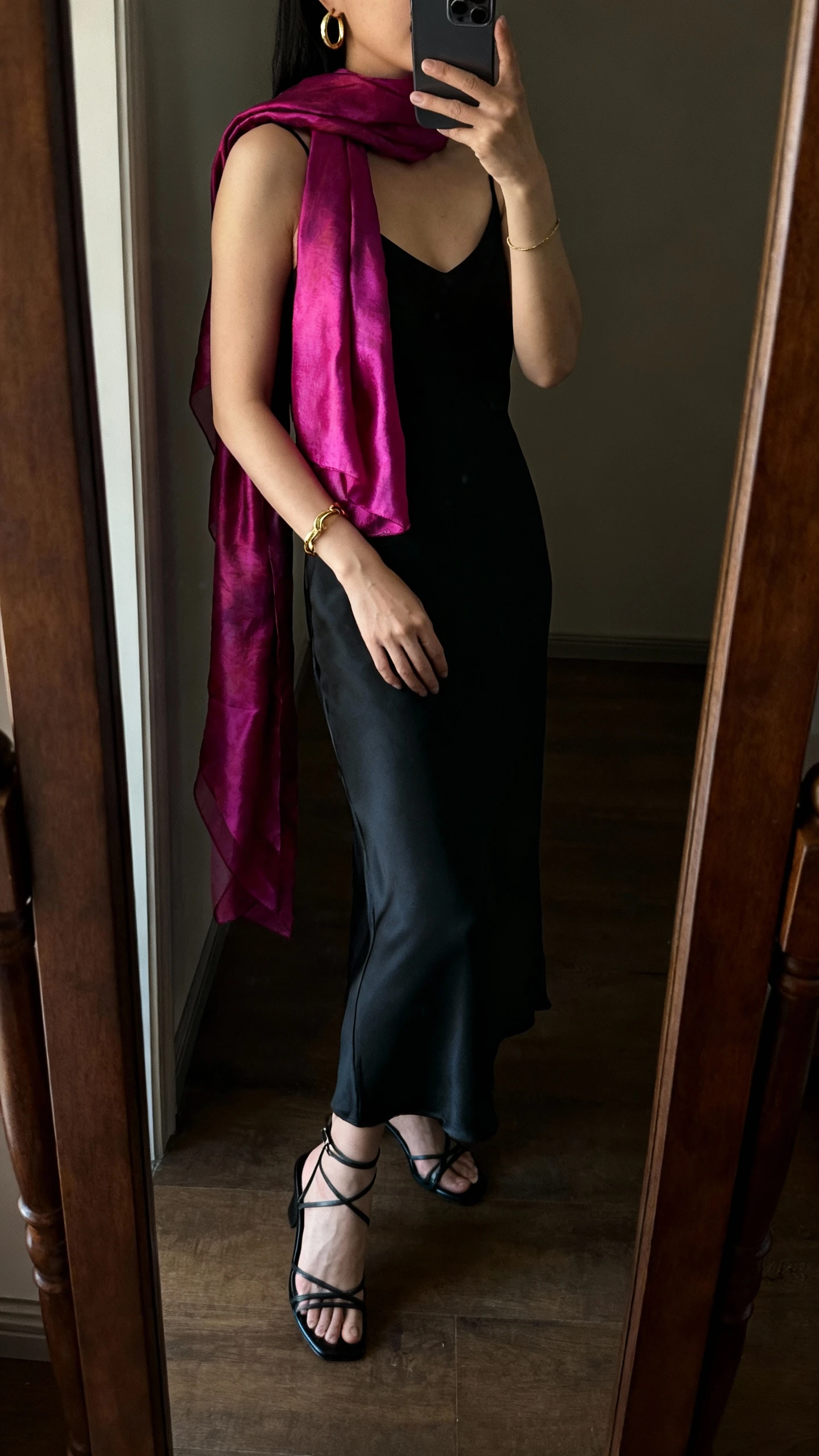 iPhone side-angle mirror selfie of a woman wearing a sleek black slip dress, strappy heels, delicate gold hoops and bracelet, and a luminous jewel-tone scarf worn as a shoulder wrap, face not visible, cropped at neck, full-length mirror in dim warm lighting, iPhone photo quality.
