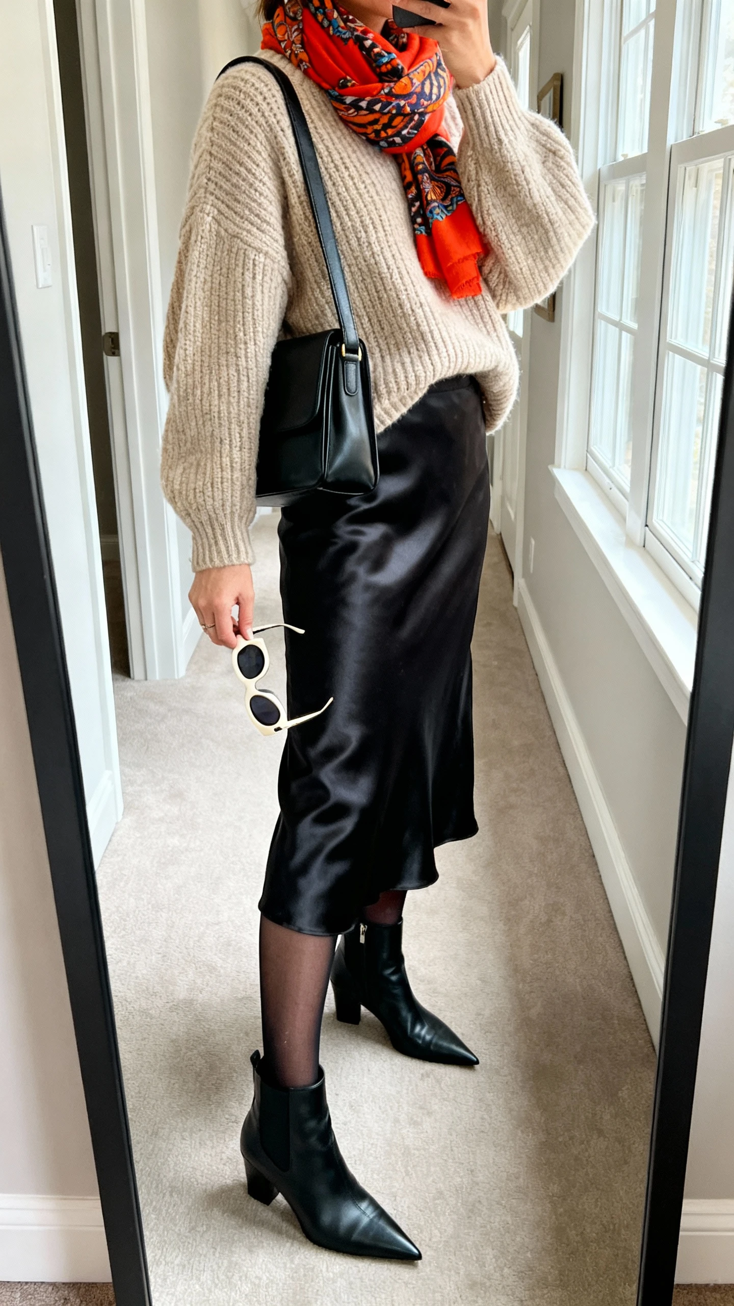 iPhone side-angle mirror selfie of a woman wearing an oversized oatmeal fisherman sweater, a black satin midi skirt, sheer black tights, pointed black ankle boots, a structured black shoulder bag, statement sunglasses in hand, and a bright patterned scarf, face not visible, cropped at shoulders, full-length mirror in hallway, soft window light, iPhone photo quality.