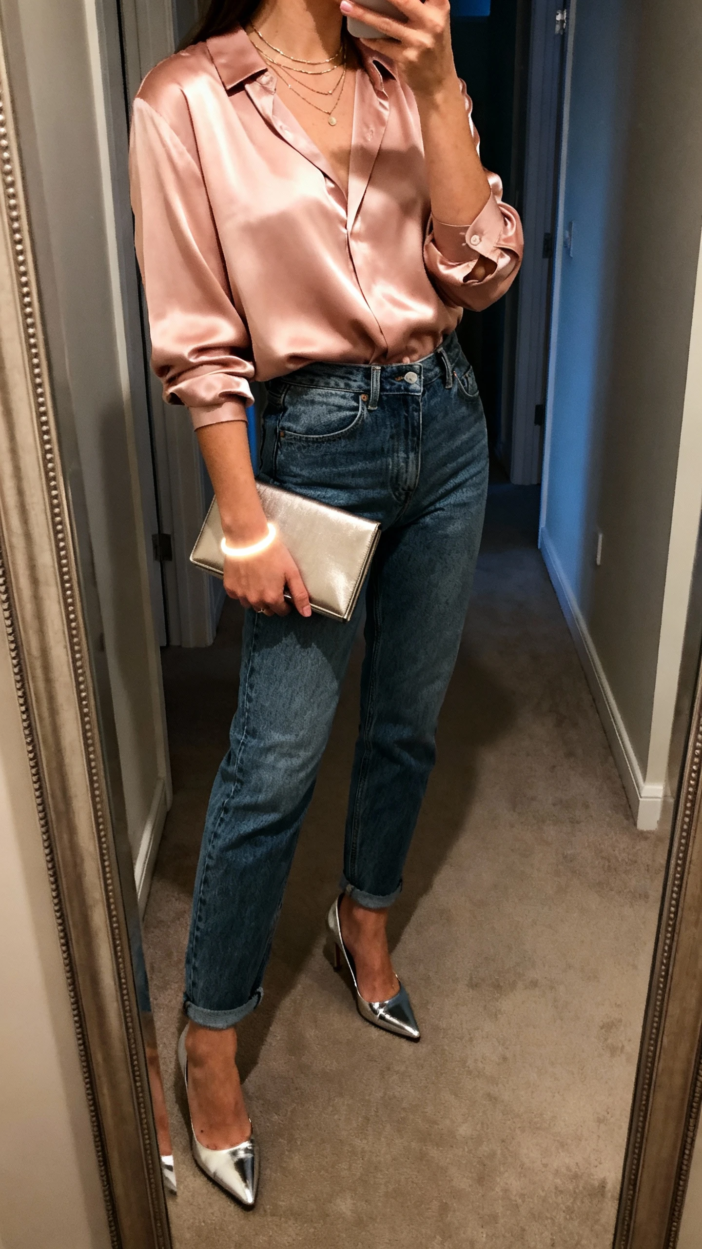 iPhone side-angle mirror selfie of a woman wearing mom jeans, a silky blush blouse tucked in, statement heels (metallic), a slim clutch, layered delicate necklaces, and a subtle glow bracelet, face not visible, hallway mirror, evening indoor light, iPhone photo quality.