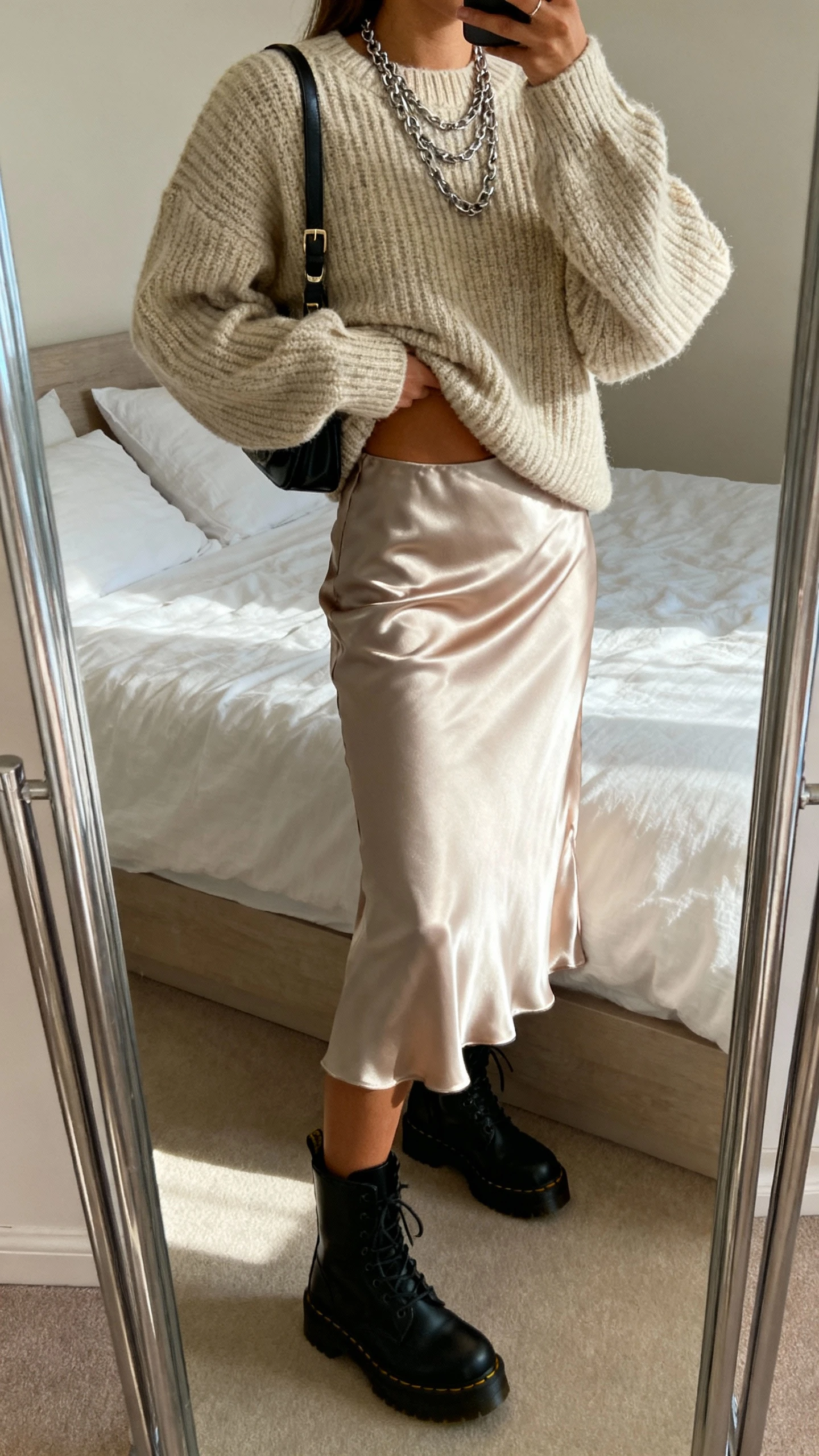 iPhone side-angle mirror selfie of a woman wearing a satin slip skirt, chunky knit sweater half-tucked, black combat boots, layered chains, and a small shoulder bag, face not visible, cropped at shoulders, bedroom mirror, natural afternoon light, iPhone photo quality.
