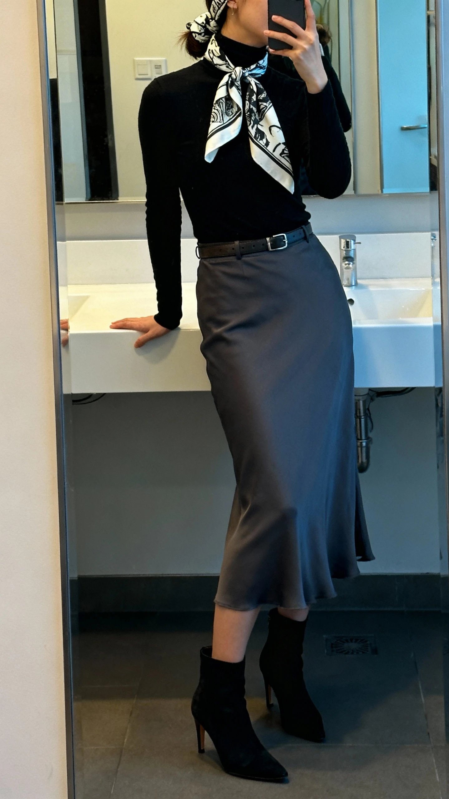 iPhone side-angle mirror selfie of a woman wearing a charcoal midi slip skirt, fitted black turtleneck, heeled ankle boots, minimalist leather belt, and a bold graphic scarf tied as a headband then re-tied at the neck, face not visible, cropped at shoulders, office bathroom mirror transitioning to evening lighting, iPhone photo quality.