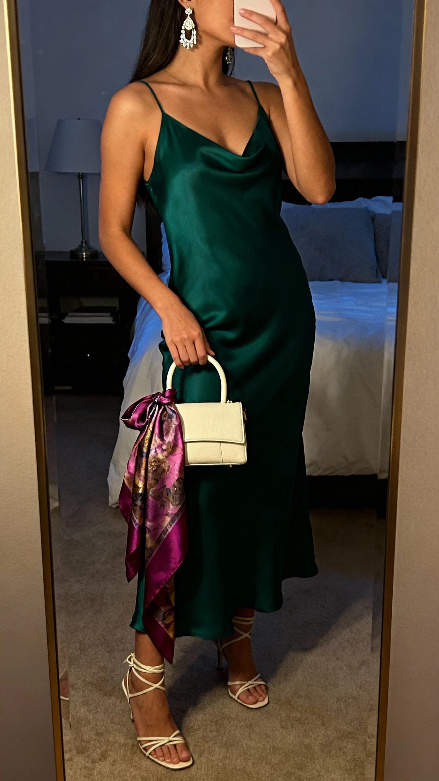 iPhone side-angle mirror selfie of a woman wearing a deep emerald slip dress, strappy heels, a mini top-handle bag with a jewel-tone silk scarf draped waterfall-style from the handle, and statement drop earrings; face not visible, bedroom mirror at dusk, warm indoor lighting, iPhone photo quality.