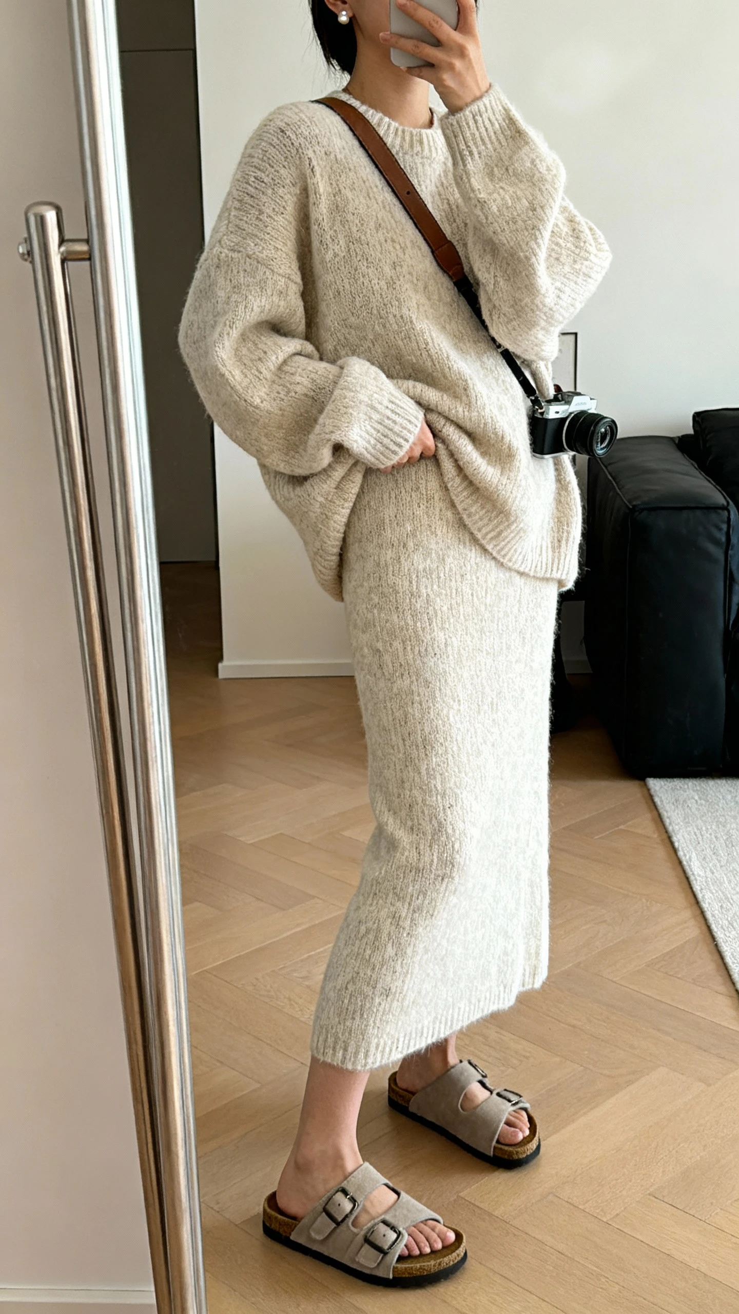 iPhone side-angle mirror selfie of a woman wearing a monochrome knit set: slouchy knit sweater and matching knit midi skirt, chunky slip-on sandals, crossbody camera bag, and subtle stud earrings, face not visible, living room full-length mirror with soft indoor lighting, iPhone photo quality.